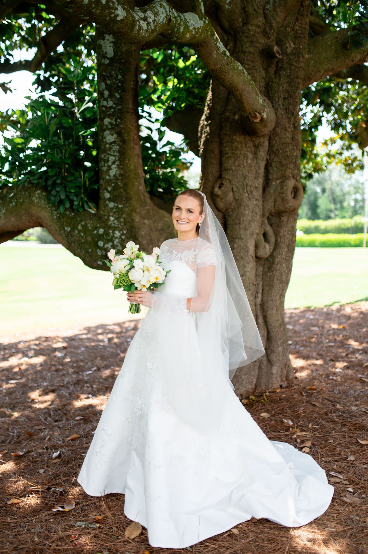 Bride in lace wedding dress holds white bouquet while posing under large oak tree with veil flowing.