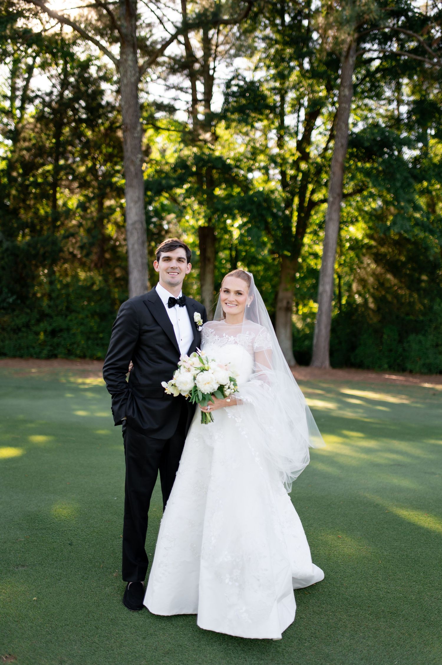 Bride and groom pose on lush green golf course, bride wearing lace gown with veil holding white bouquet.