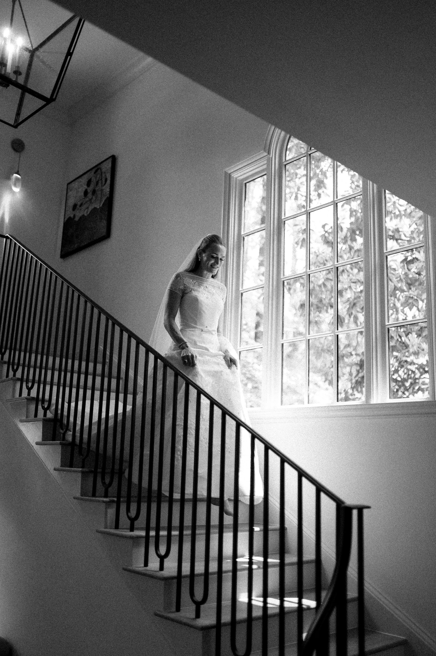 Bride descending grand staircase in black and white wedding photo with arched window backdrop.