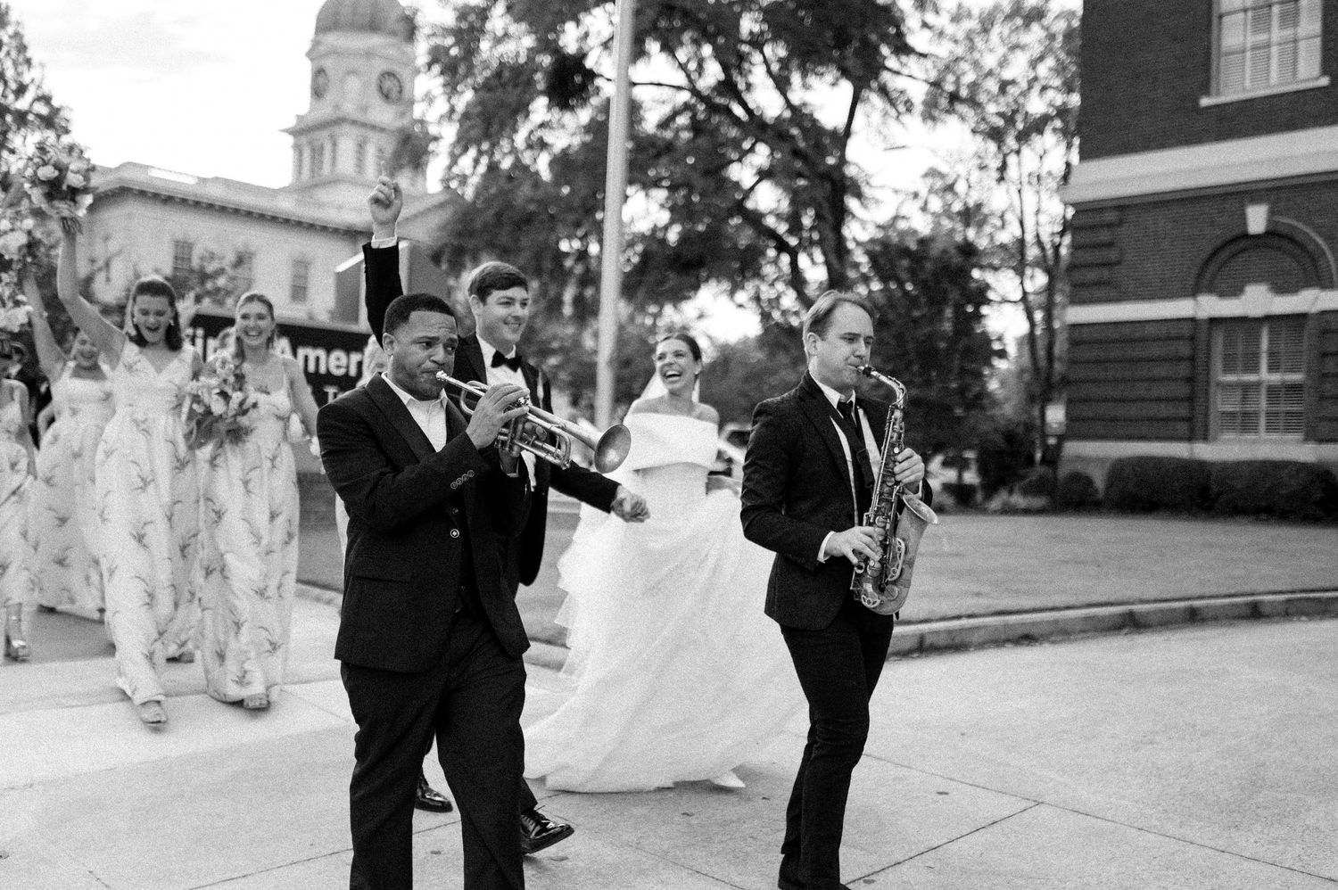 Black and white wedding processional with brass band playing trumpet and saxophone leading bride outdoors.