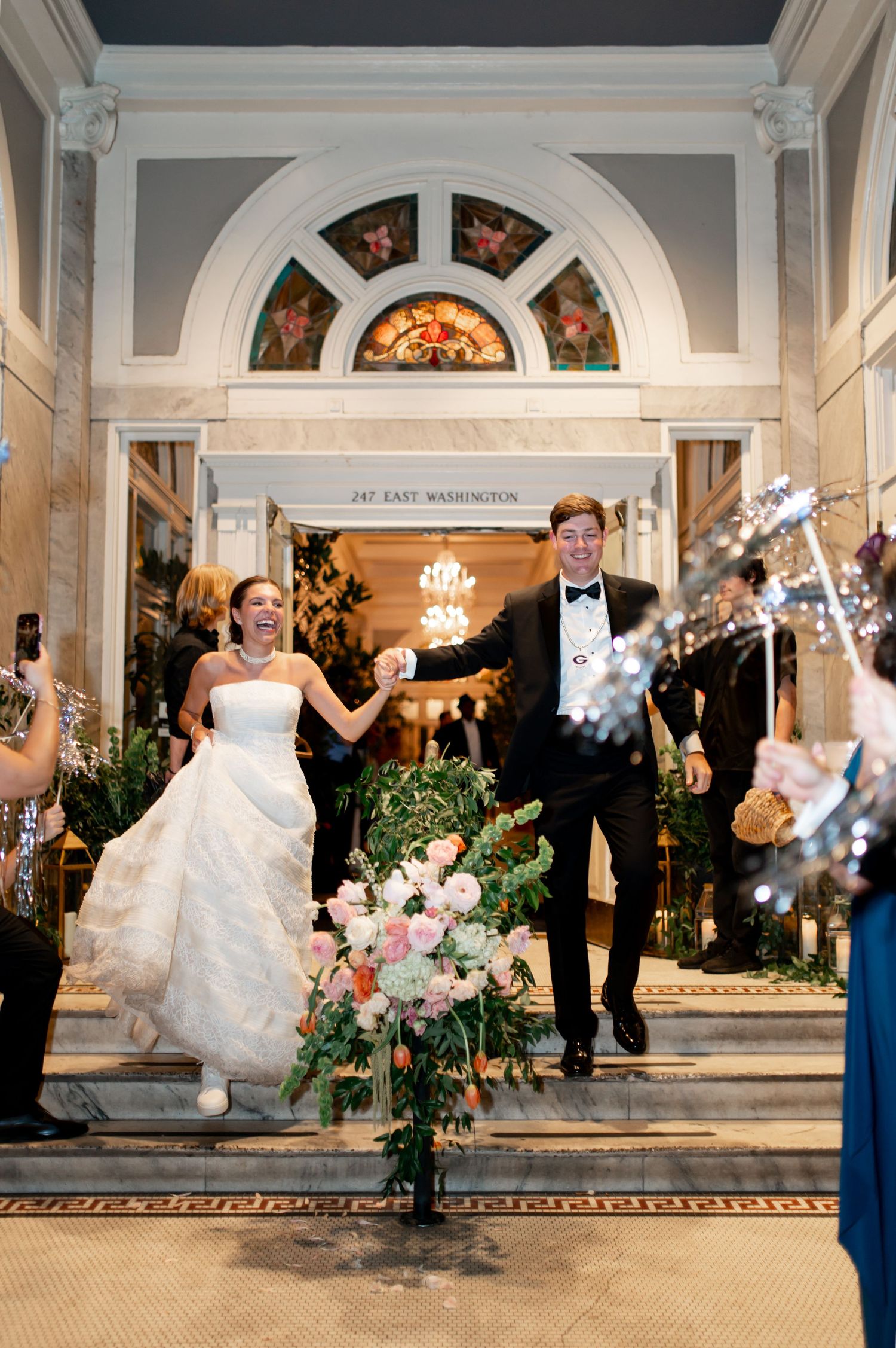 Newlyweds exit venue through grand marble foyer as guests celebrate with sparklers and cheers.