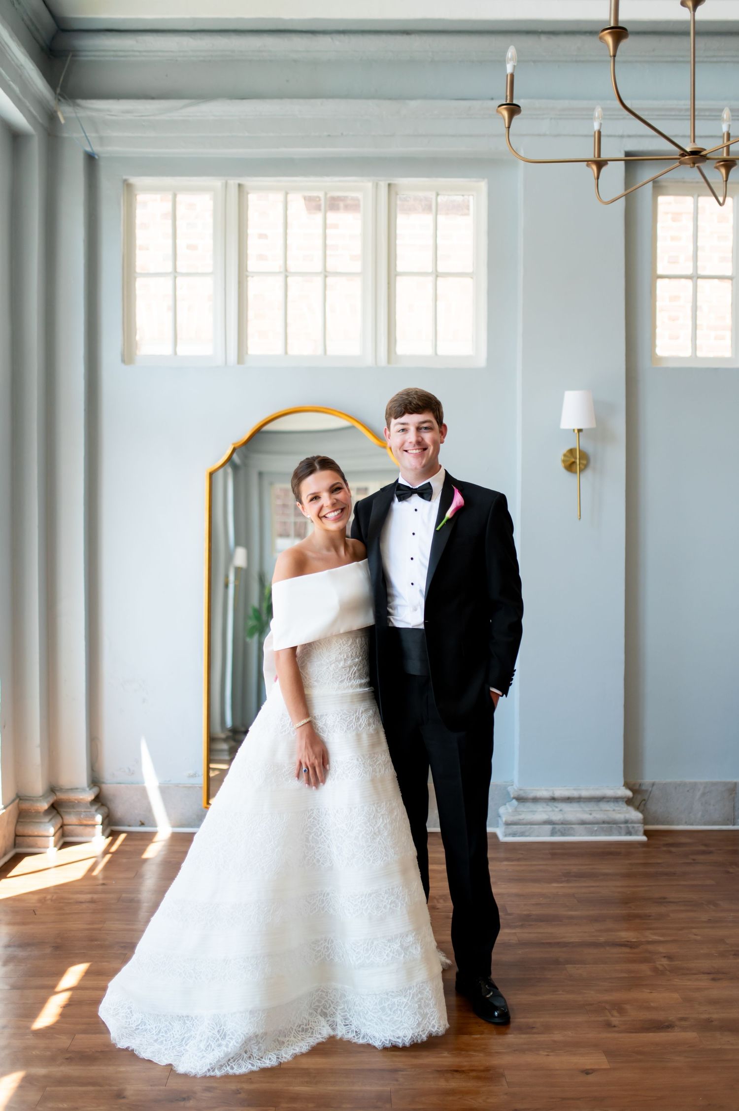 A smiling bride in an off-shoulder white gown and groom in black tuxedo pose together in an elegant blue room.