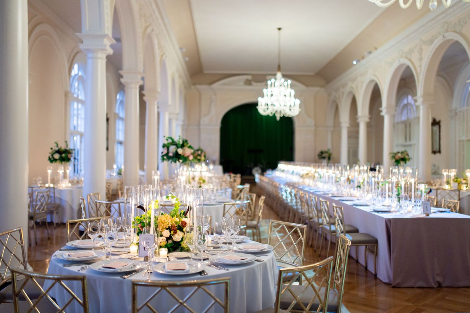 Wedding reception tables arranged in an ornate white ballroom with sparkling chandeliers.