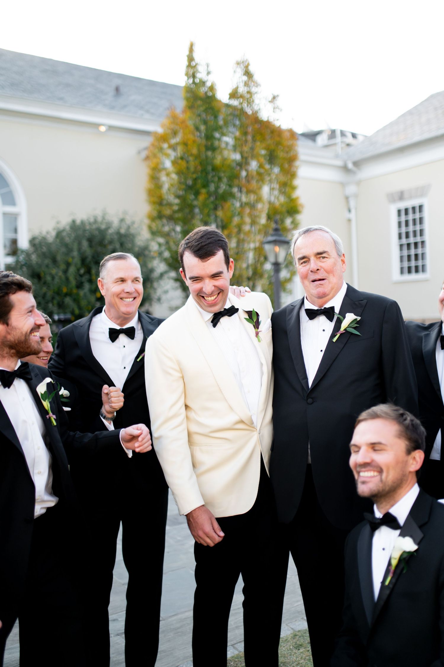 Groomsmen in black tuxedos and white dinner jacket share laughs during formal photos.