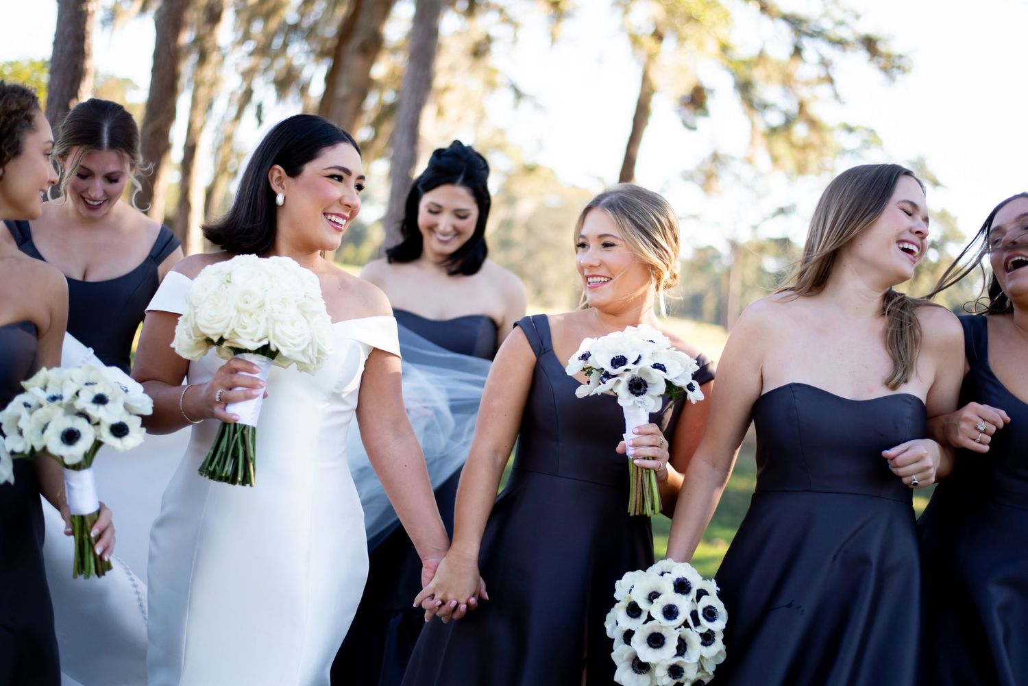 Bridal party in navy dresses and white gown pose with bouquets under trees.