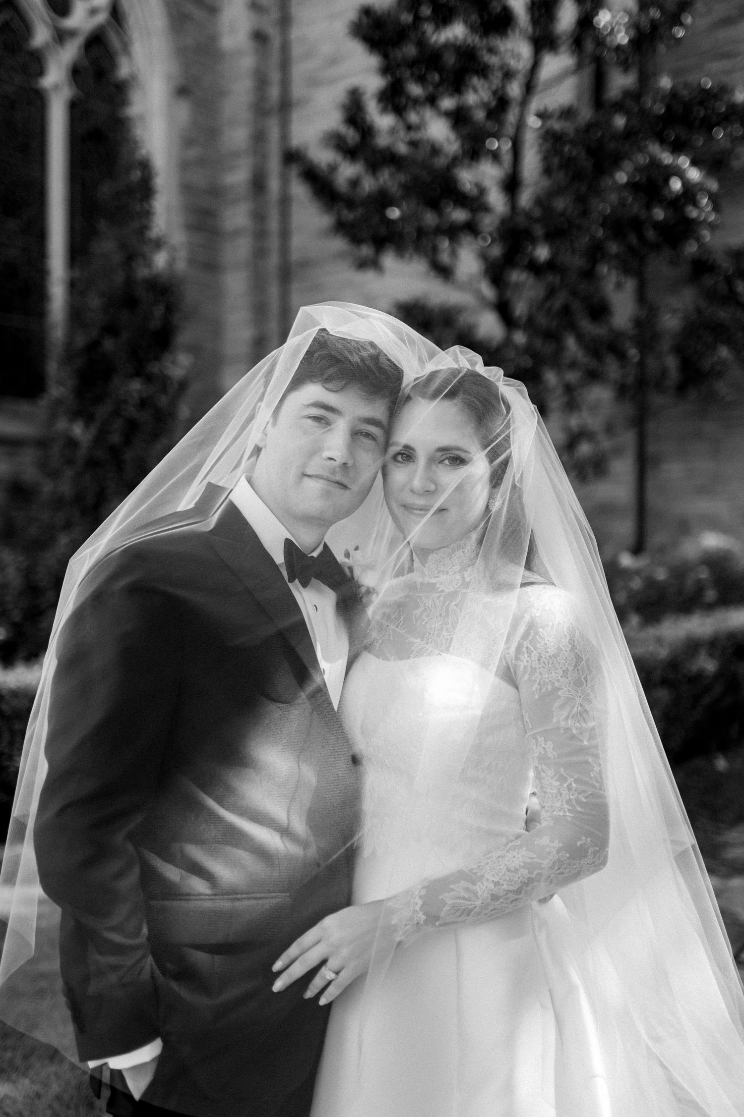 Black and white wedding portrait of bride in lace gown and veil with groom in tuxedo posing outdoors.