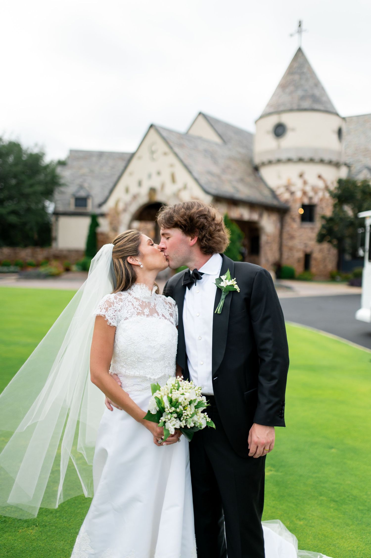 A wedding couple shares a romantic kiss on the lawn in front of a stone church with a spire.