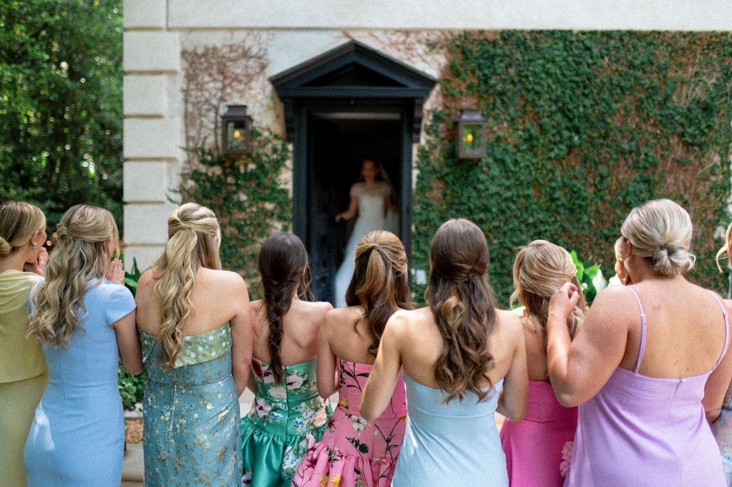 Bridesmaids in colorful dresses view bride's reveal at ivy-covered venue entrance.