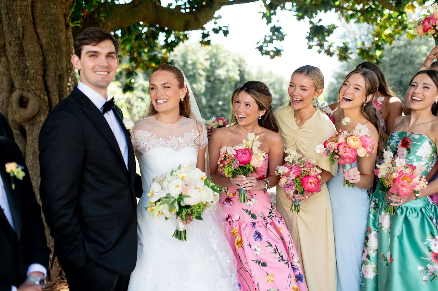 Bride and groom smile with bridesmaids in colorful floral and pastel gowns outdoors.