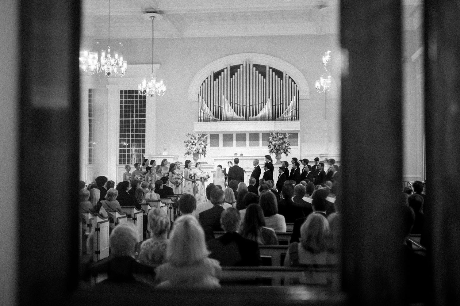 Black and white photo of a wedding ceremony inside a classic church with pipe organ and chandelier lighting.