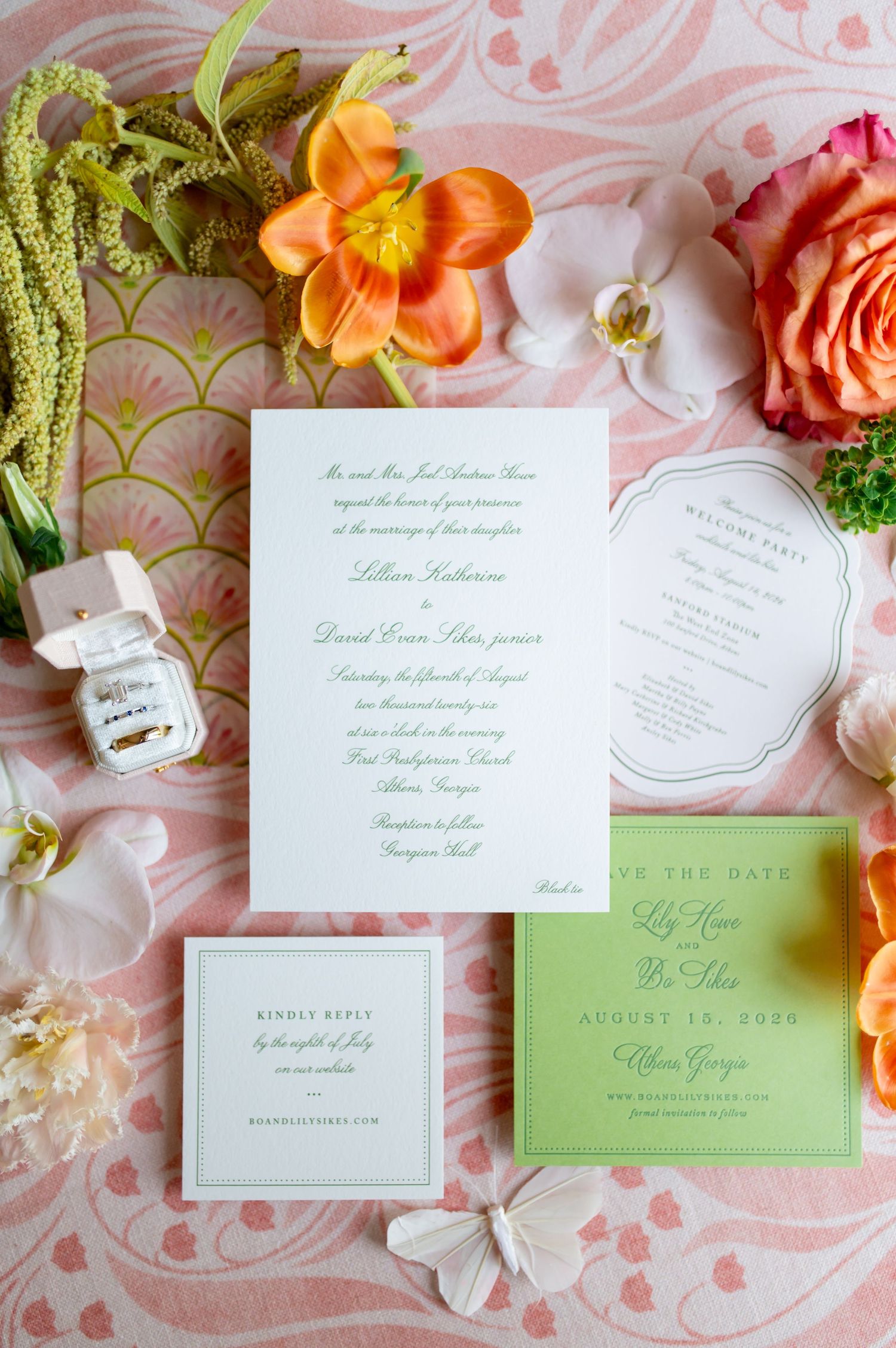 Elegant wedding invitation suite with green script on white cards, surrounded by orange lilies and pink roses on a pink floral backdrop.
