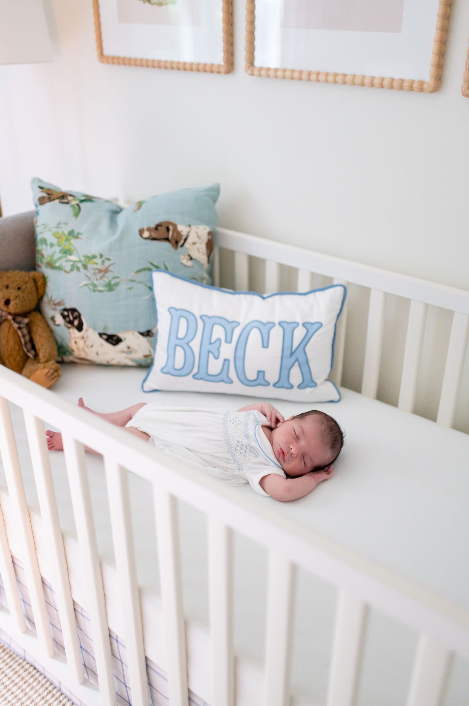 A sleeping newborn in a white crib with decorative pillows and a personalized blue name cushion.