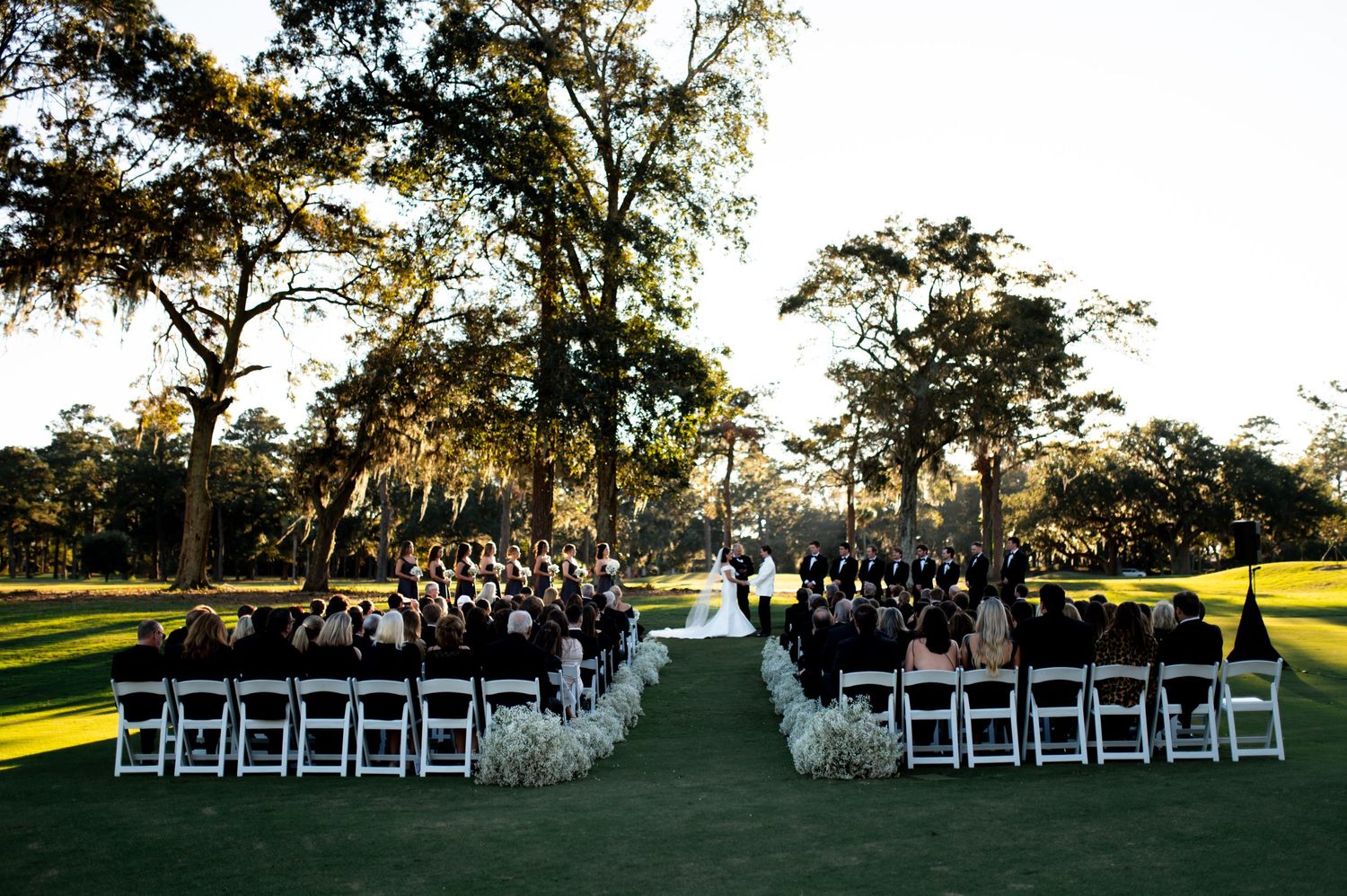 Wide view of a wedding ceremony setup with white chairs arranged on a rolling lawn beneath oak trees.