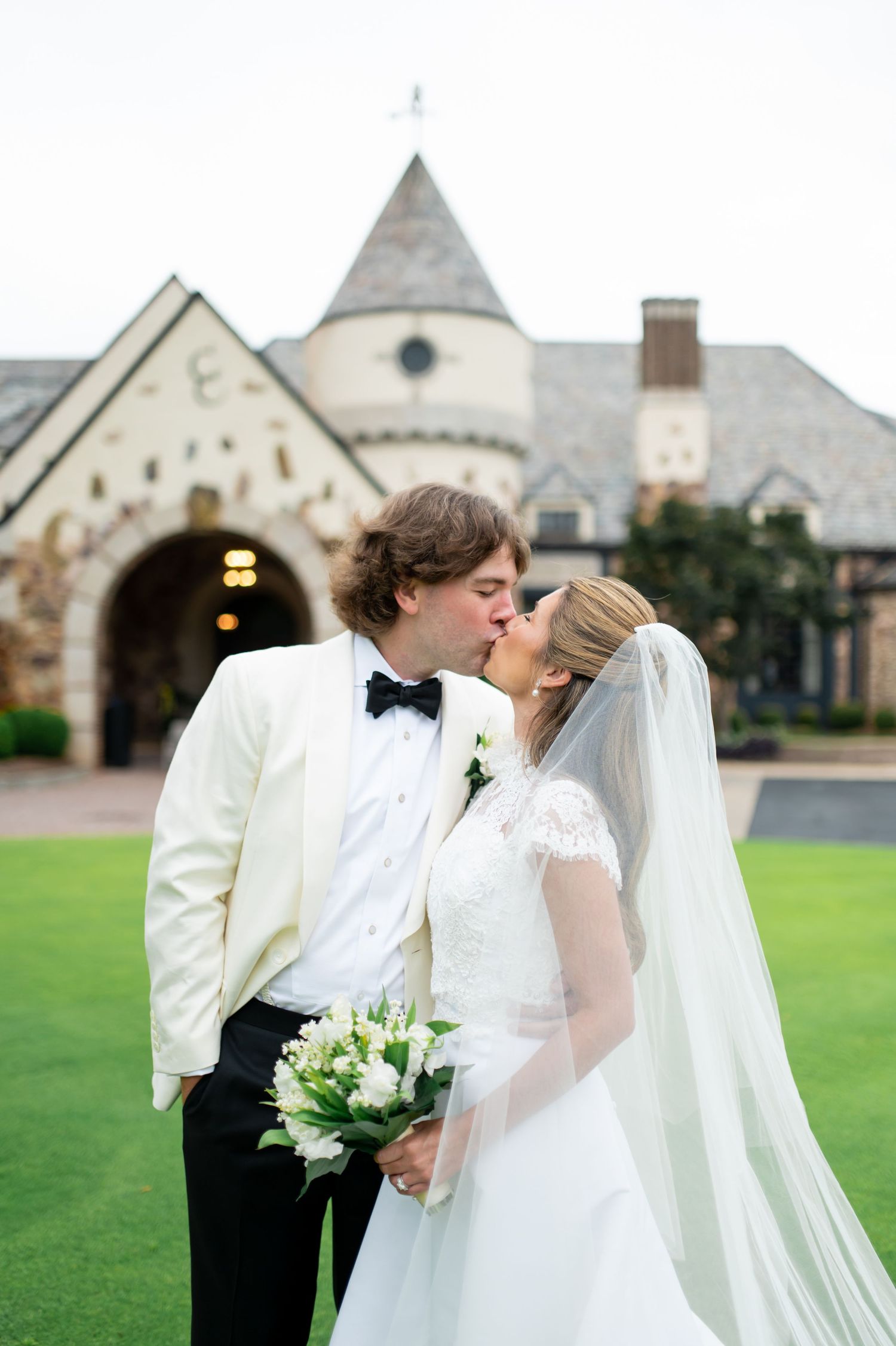 Romantic kiss between newlyweds outside stone church with white bouquet and flowing veil.