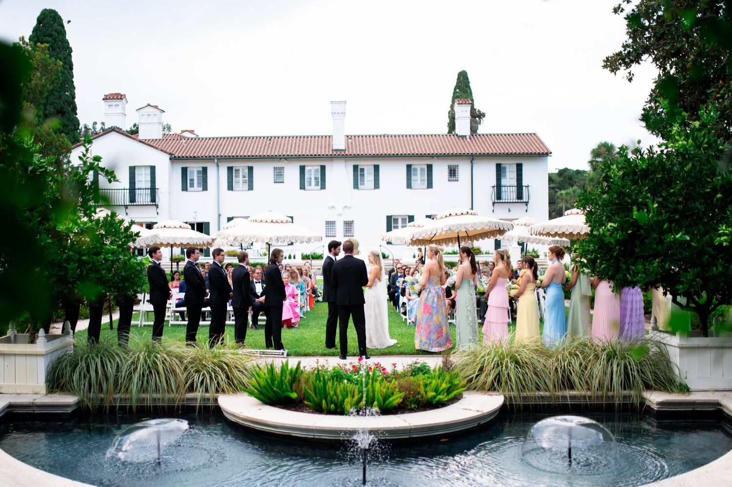 Panoramic view of an elegant white mansion with manicured gardens and a reflecting pool.
