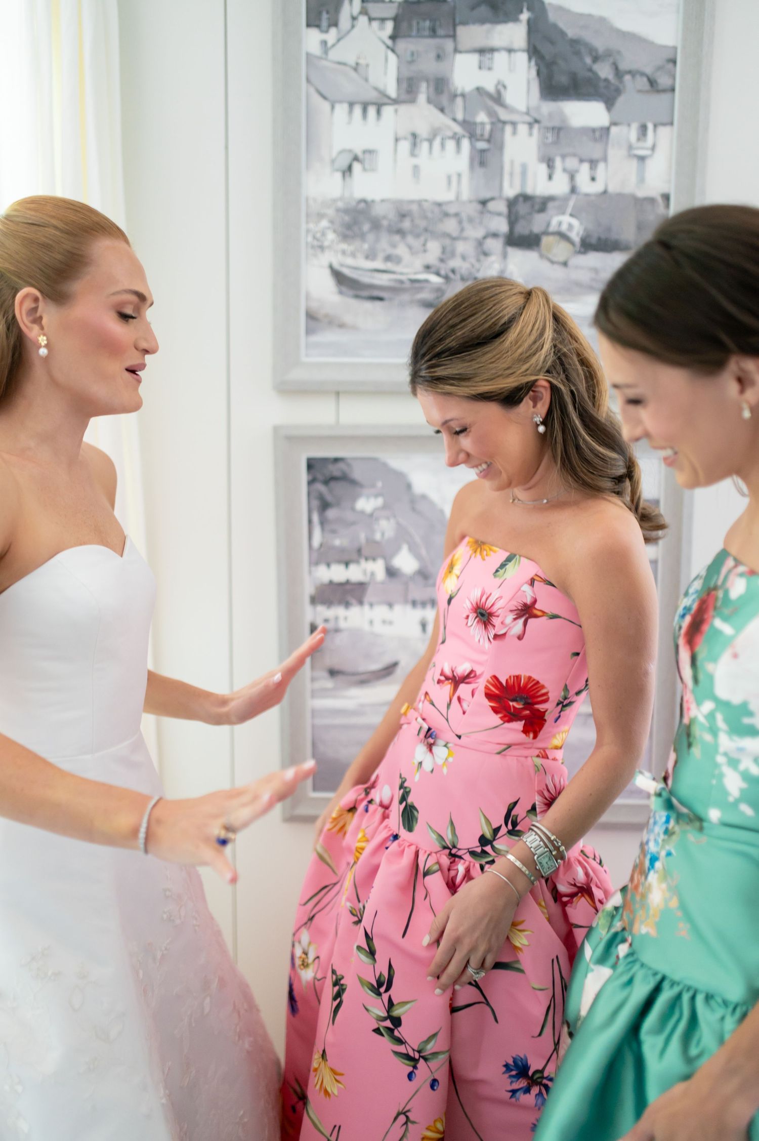 Bride in white gown gestures to guests in floral dresses during wedding preparations indoors.
