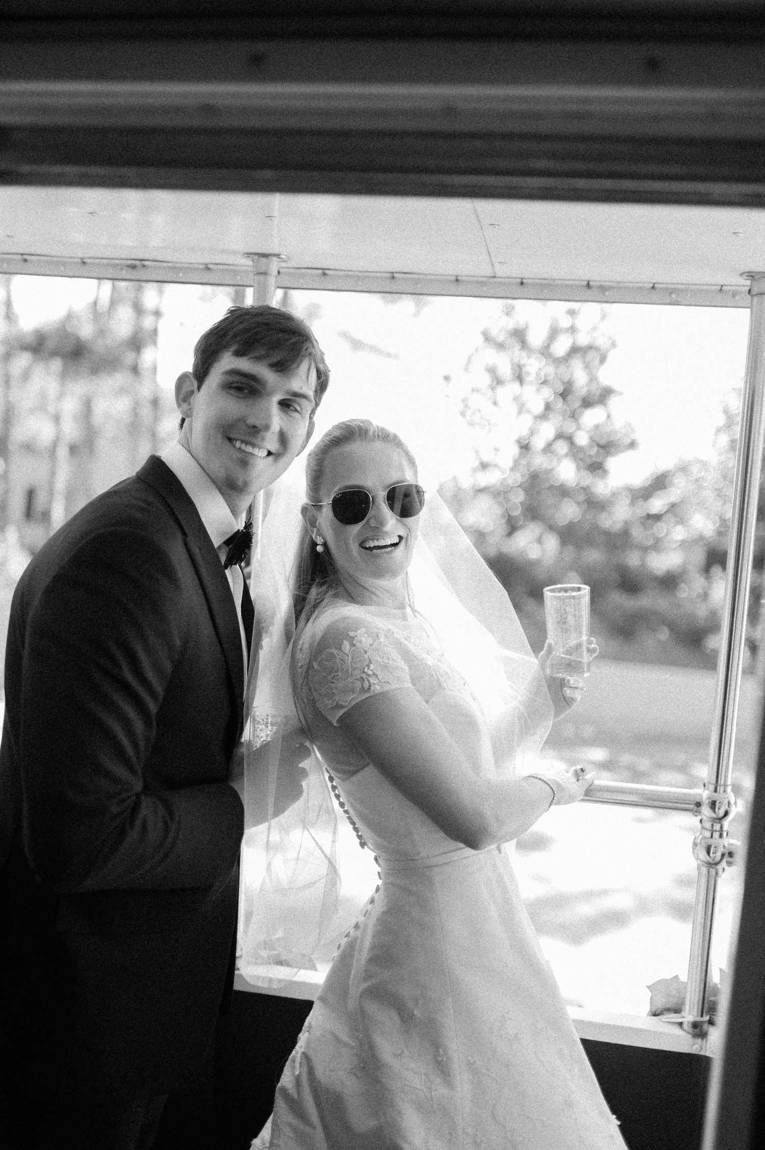 Black and white photo of smiling bride in sunglasses and groom posing together at wedding reception.