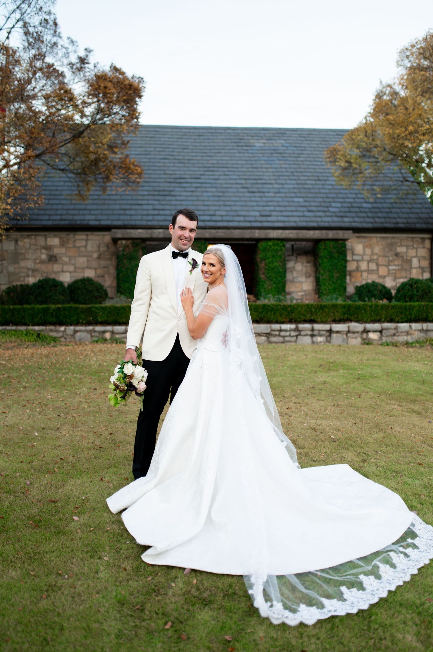 Wedding couple posing in flowing white dress against rustic stone building and autumn landscape.