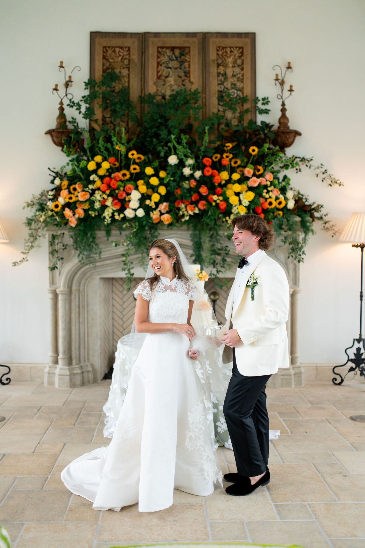 A wedding couple poses in front of an elegant stone archway decorated with vibrant orange and yellow floral arrangements.