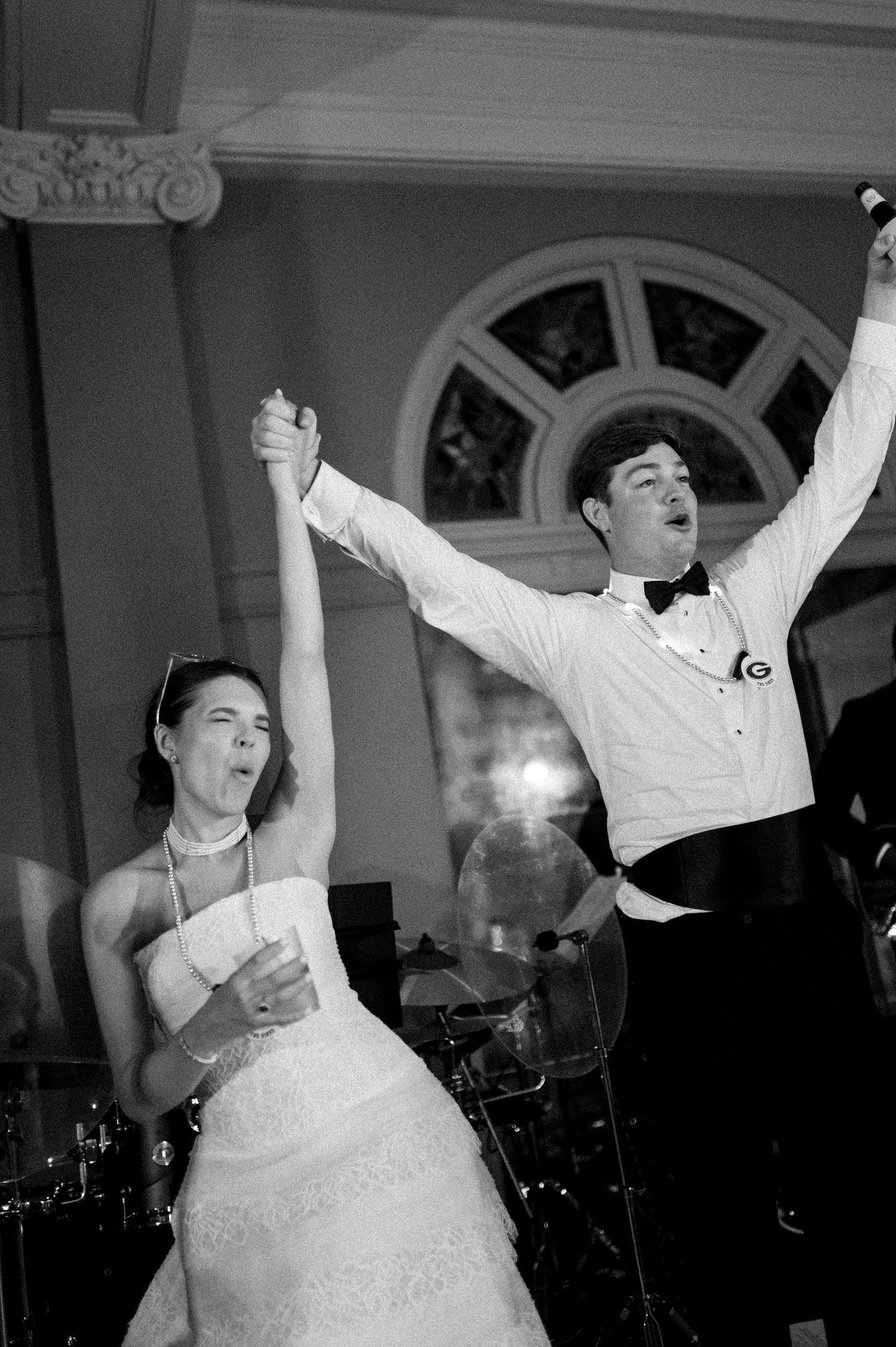 Black and white photo of bride and groom triumphantly raising arms while celebrating on wedding reception dance floor.