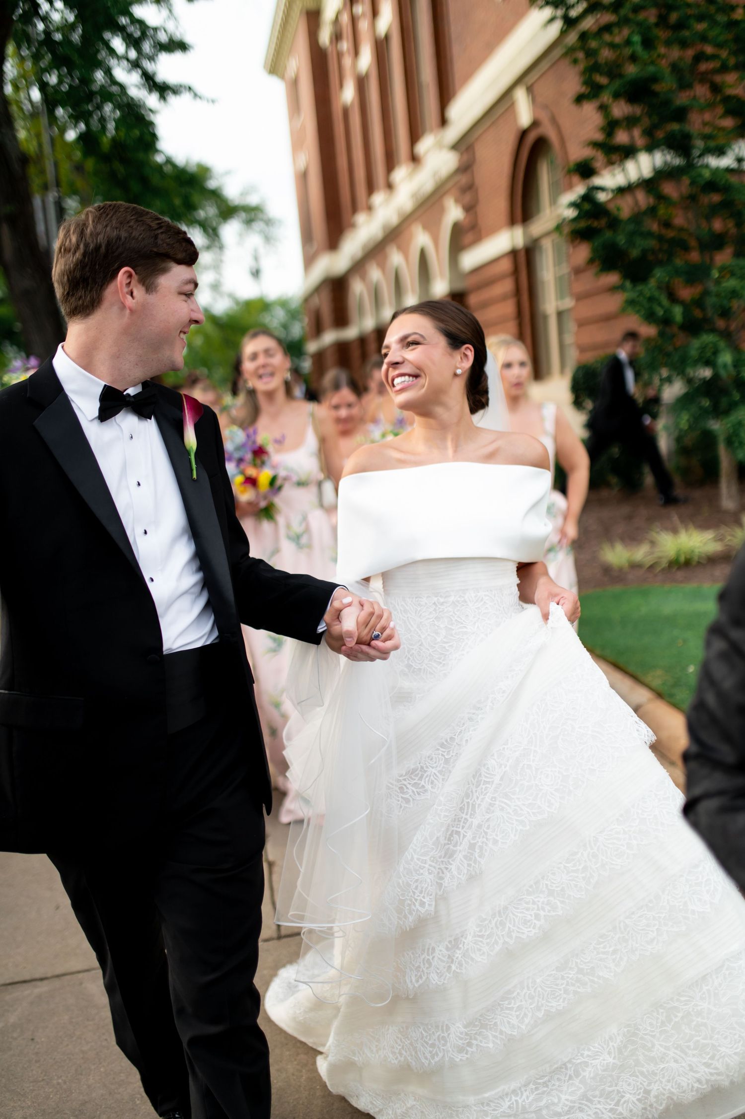 Smiling bride in off-shoulder lace gown holds grooms hand walking outdoors with wedding party.