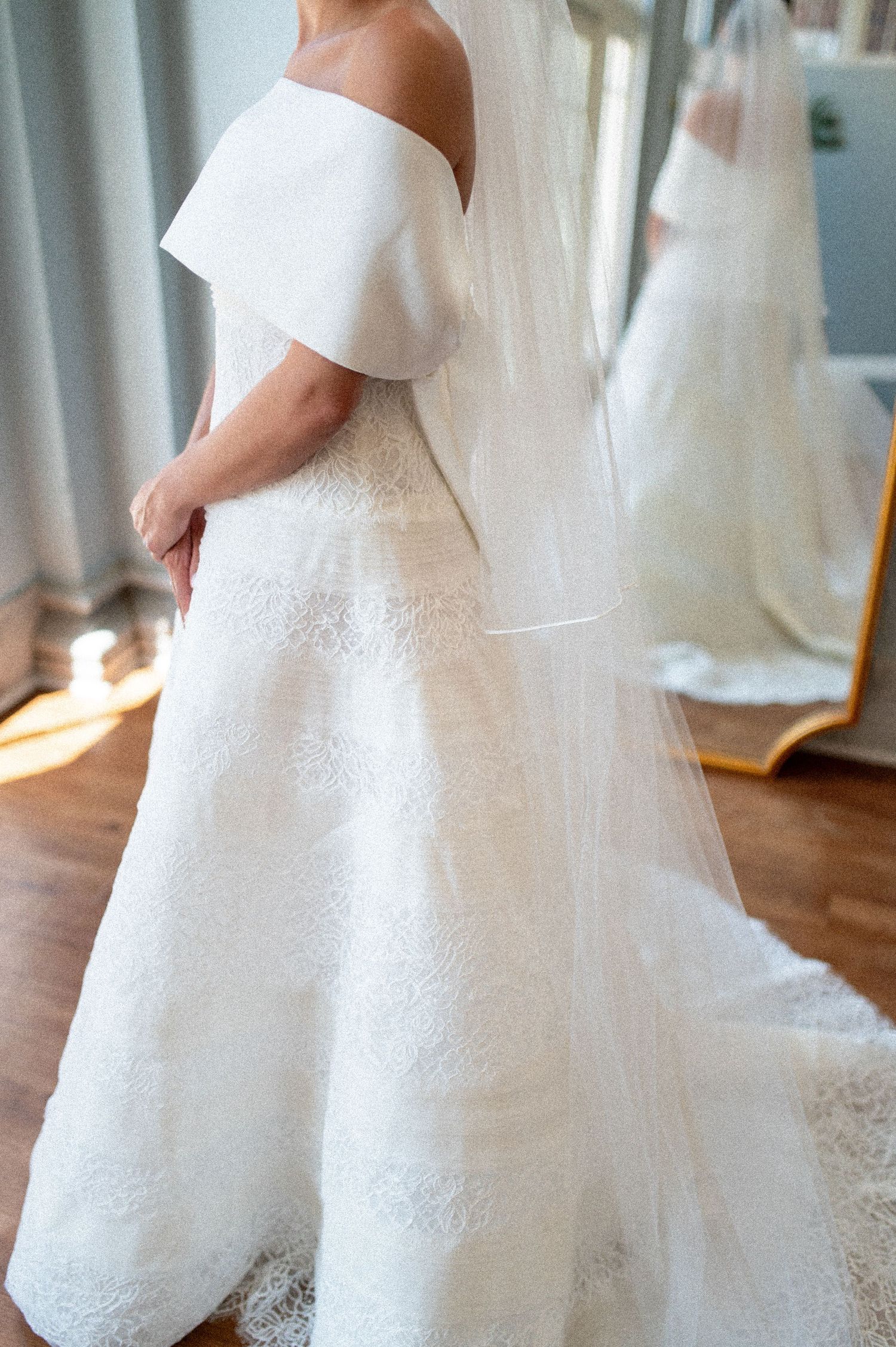 Bride wearing an off-shoulder white lace wedding gown with a flowing veil, viewed from behind in a bridal boutique.