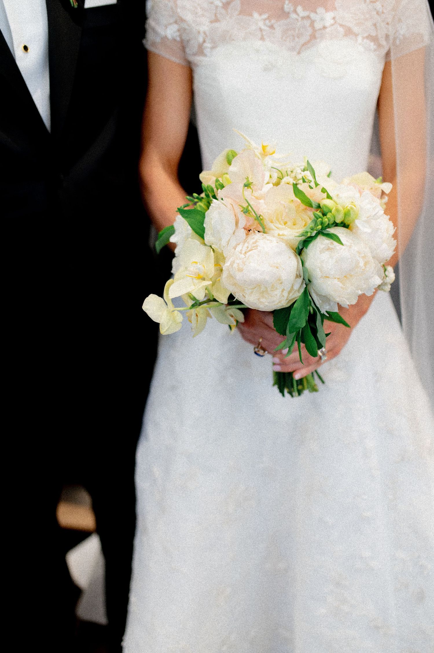 Bride holding elegant white peony bouquet with green accents against lace wedding gown.