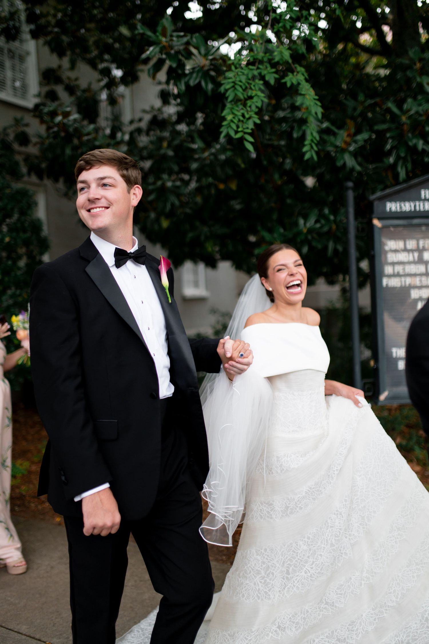 Joyful bride and groom in black tuxedo walking outside church after wedding ceremony, bride in off-shoulder gown.