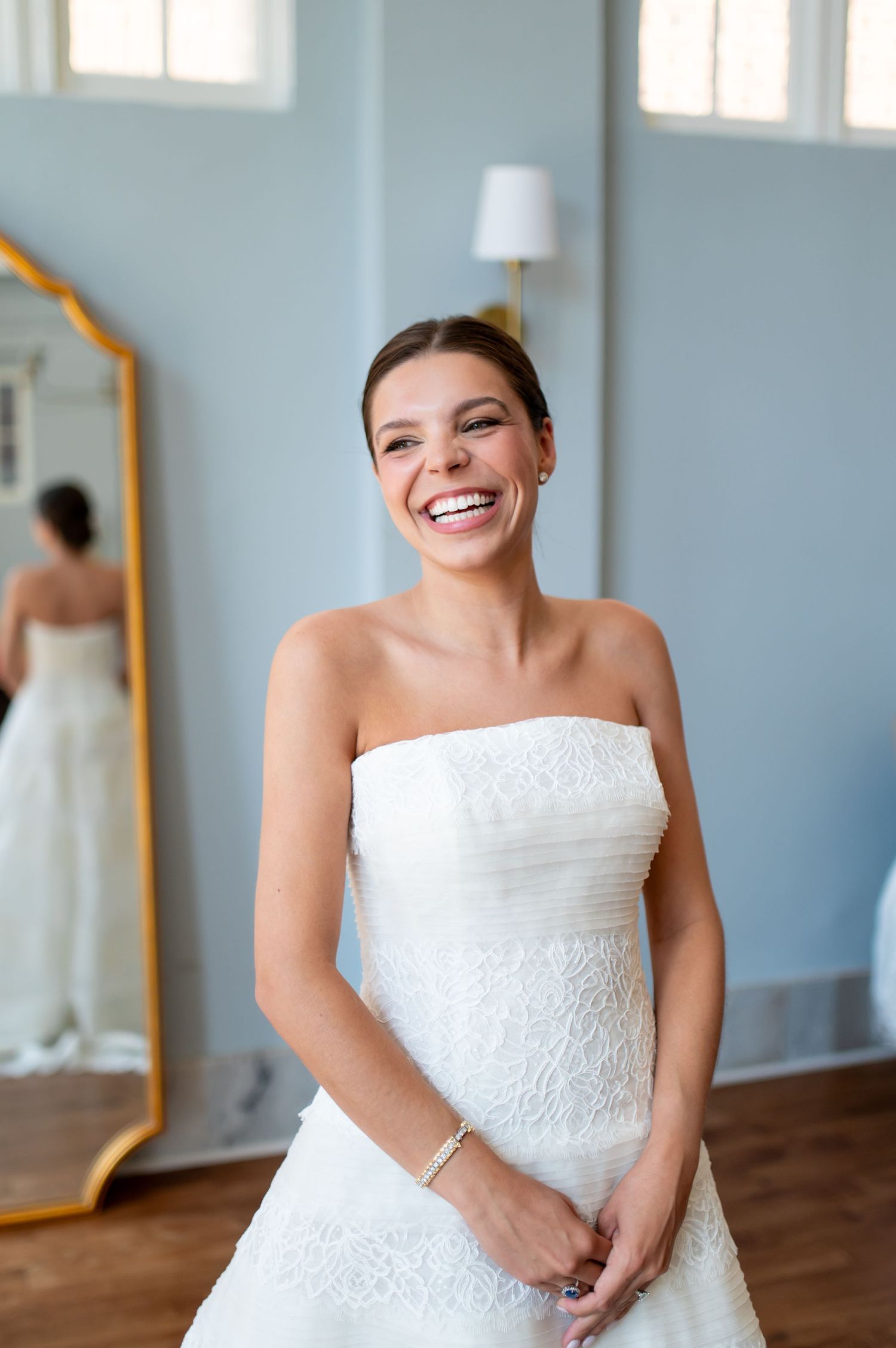 Smiling bride in strapless white lace wedding gown standing in blue room with gold mirror reflection.