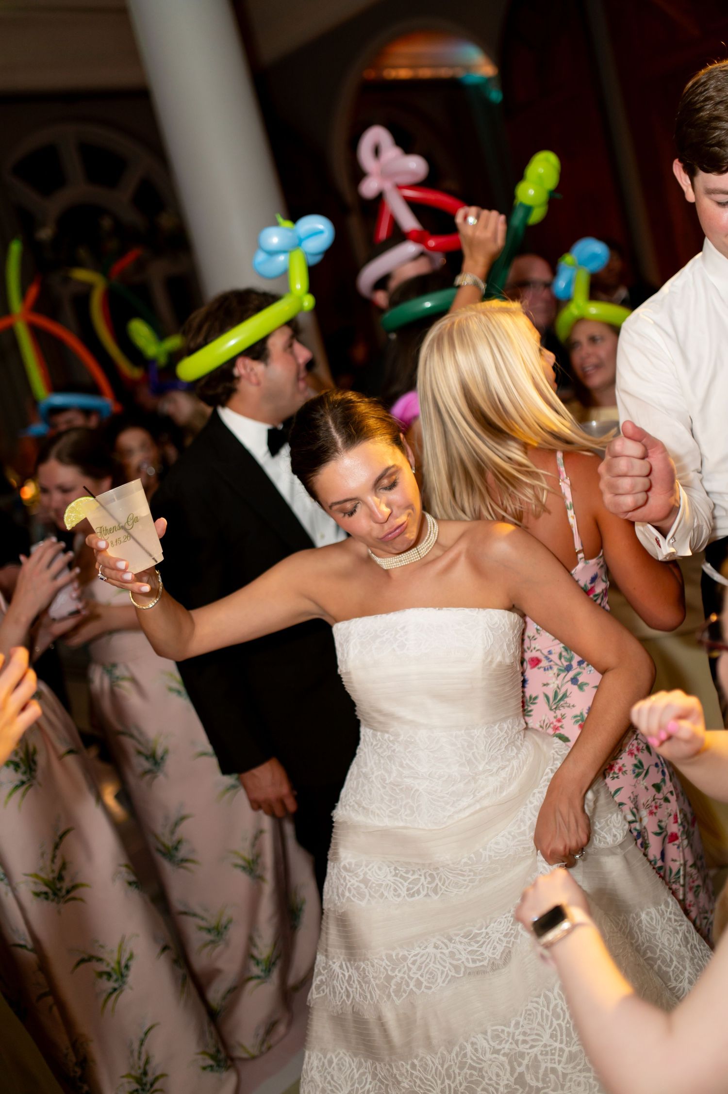 Bride in strapless lace gown dances joyfully with guests wearing balloon hats at wedding reception.