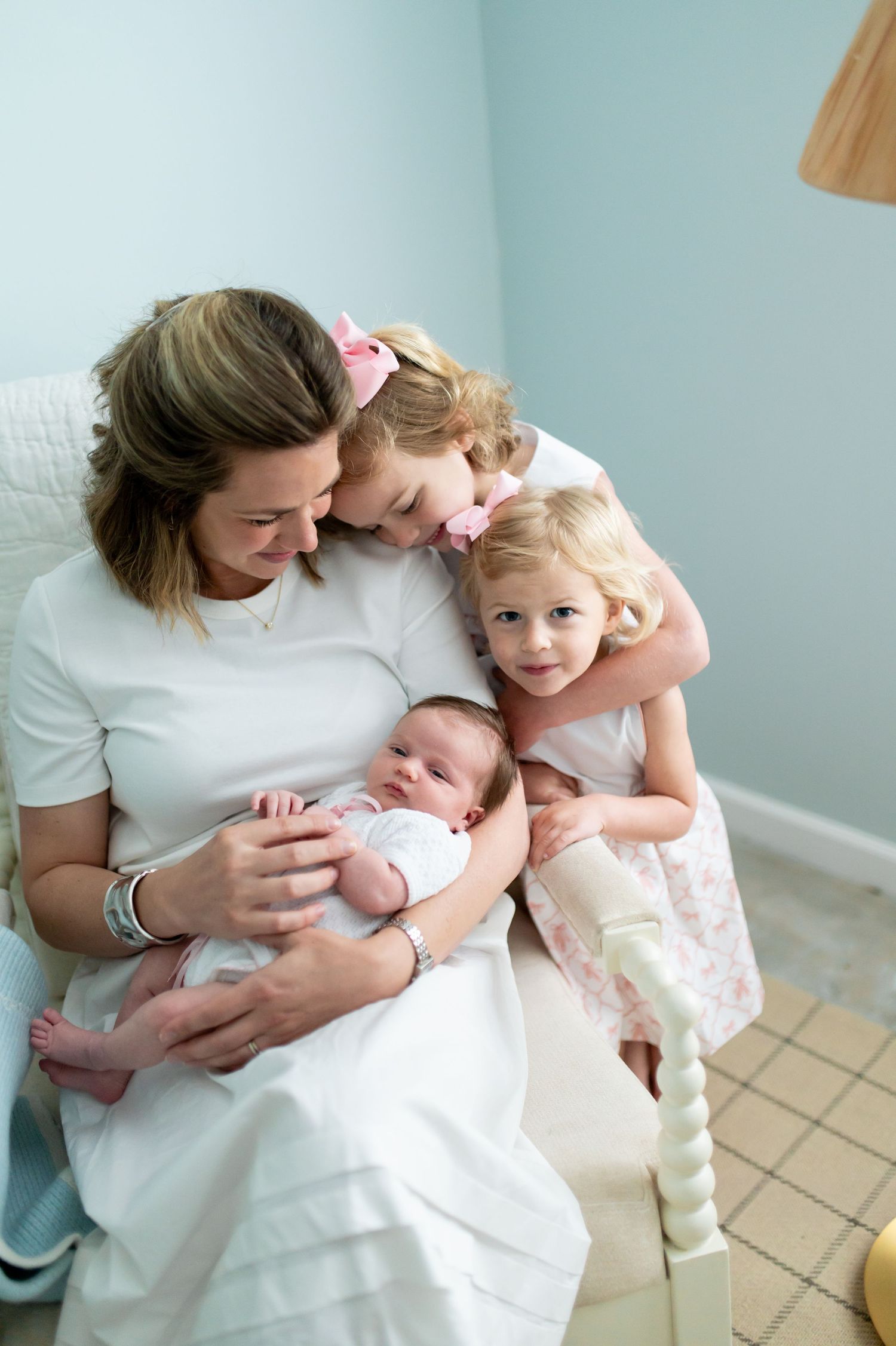 A mother sits in a chair holding a newborn while two young daughters in pink dresses lean in for a family portrait.