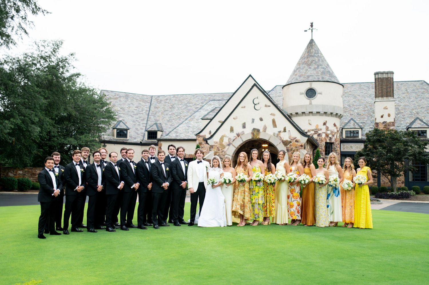 Wedding party lineup with bridesmaids in yellow and groomsmen in black against stone church backdrop.
