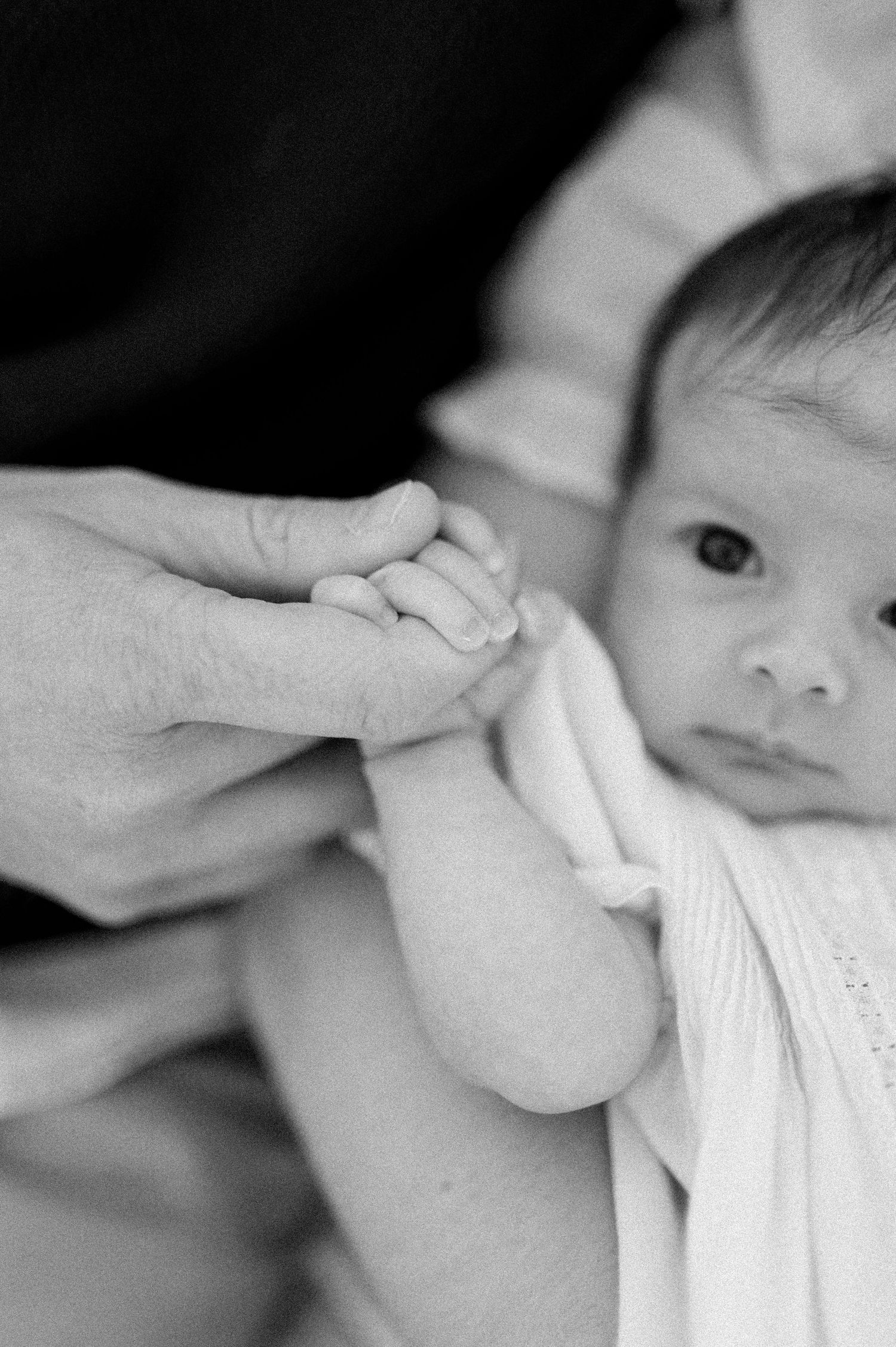 Black and white close-up of a baby's face with an adult hand gently holding their tiny hand.