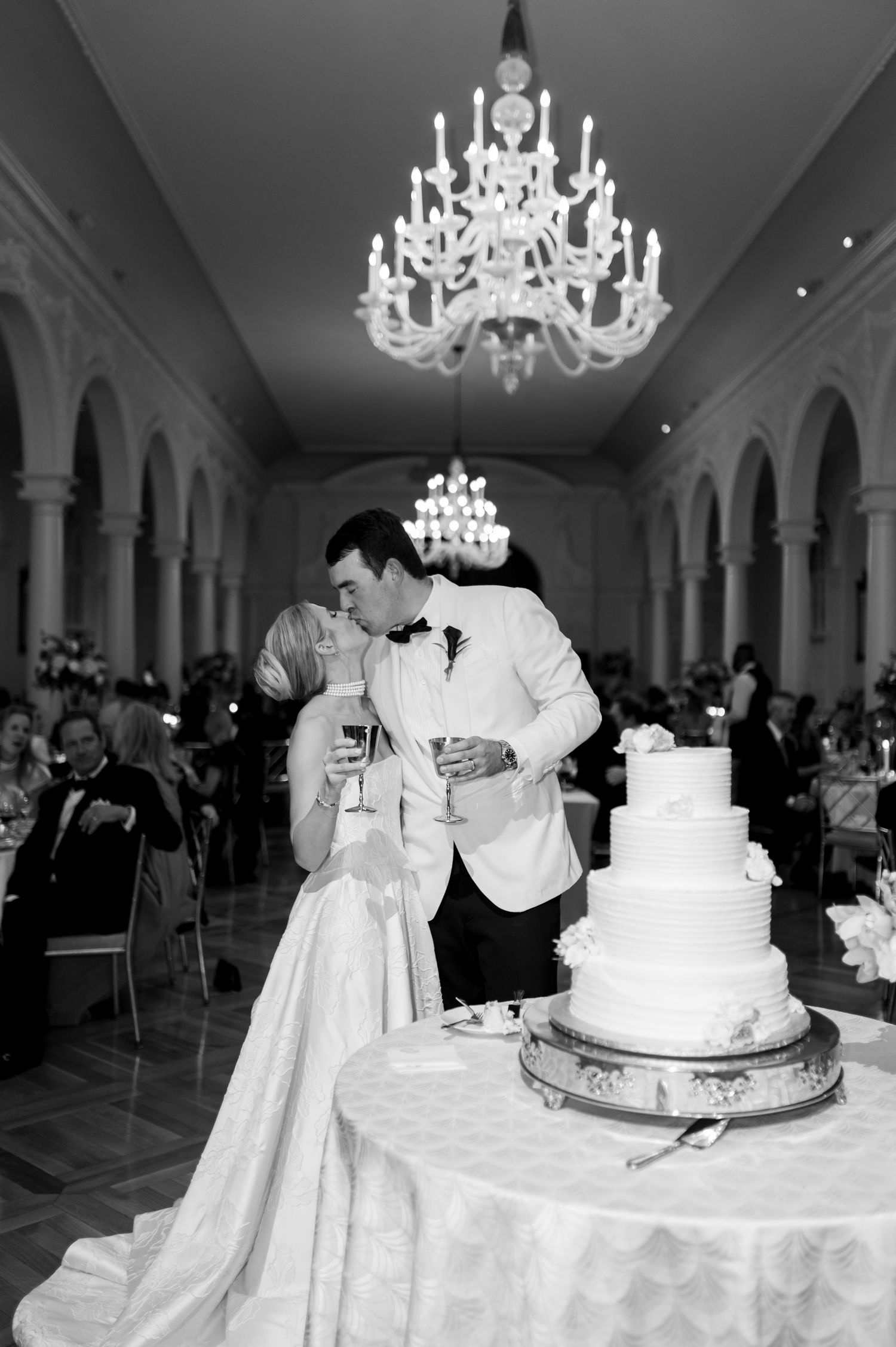 Black and white photo of newlyweds cutting their multi-tiered wedding cake beneath crystal chandeliers.