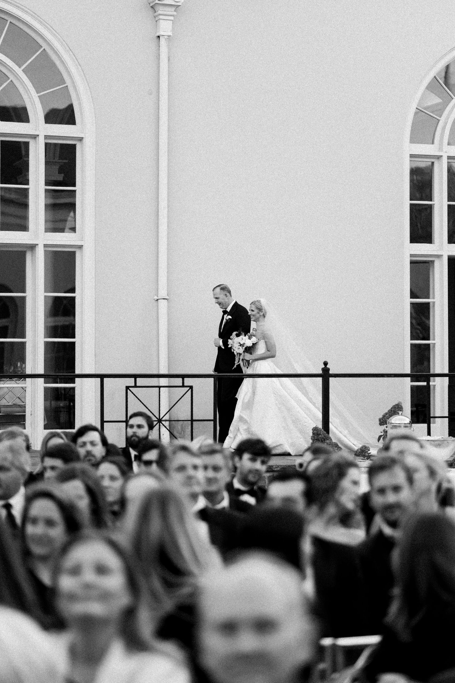 Wedding guests gather outdoors at an elegant venue with arched windows and white columns.