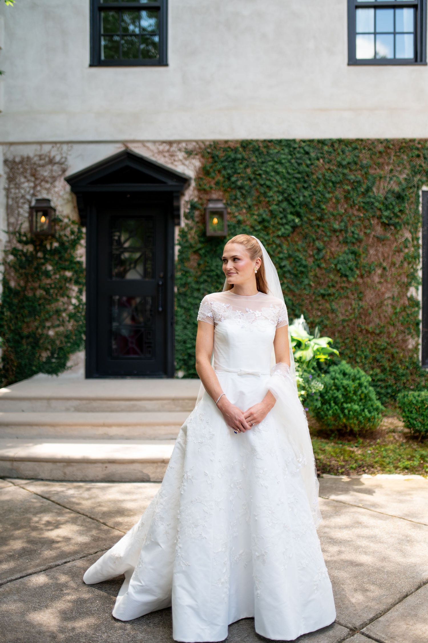 Elegant bride in a white lace ballgown and veil posing outside an ivy-covered building.