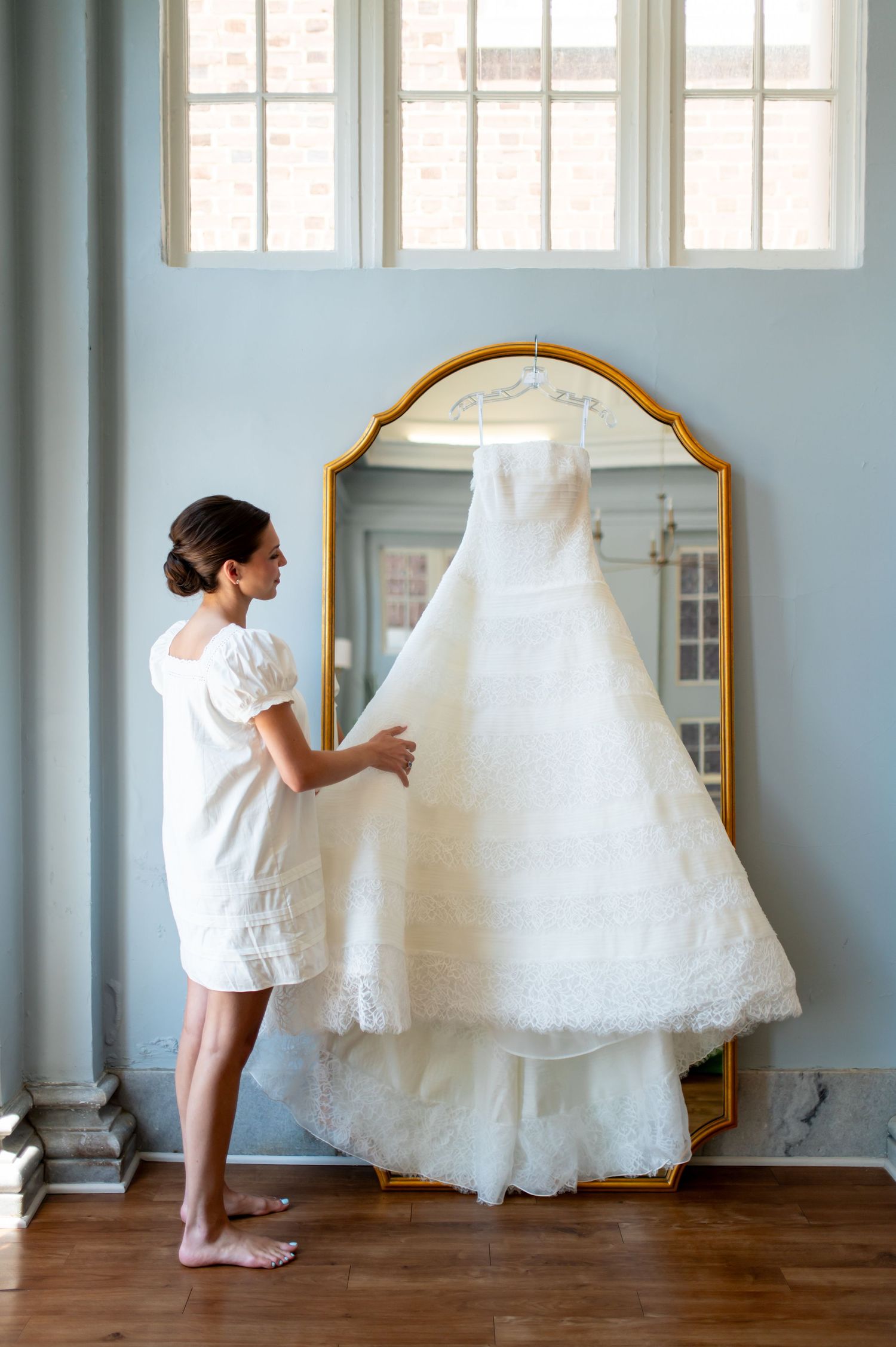 Bride in white mini dress admires her lace wedding gown hanging on a gold ornate mirror in a blue room.