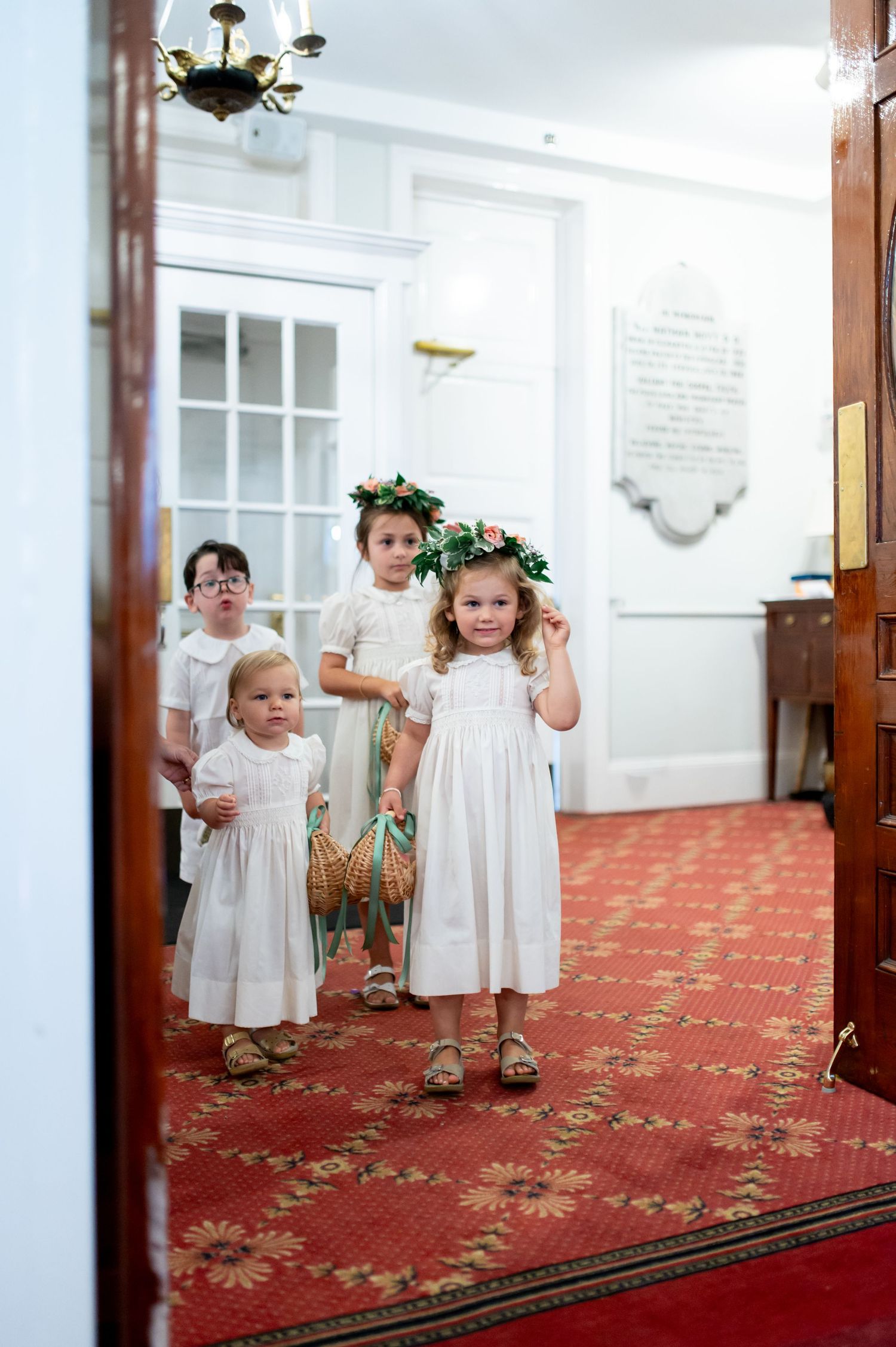 Young flower girls in white dresses with floral crowns stand in church doorway.