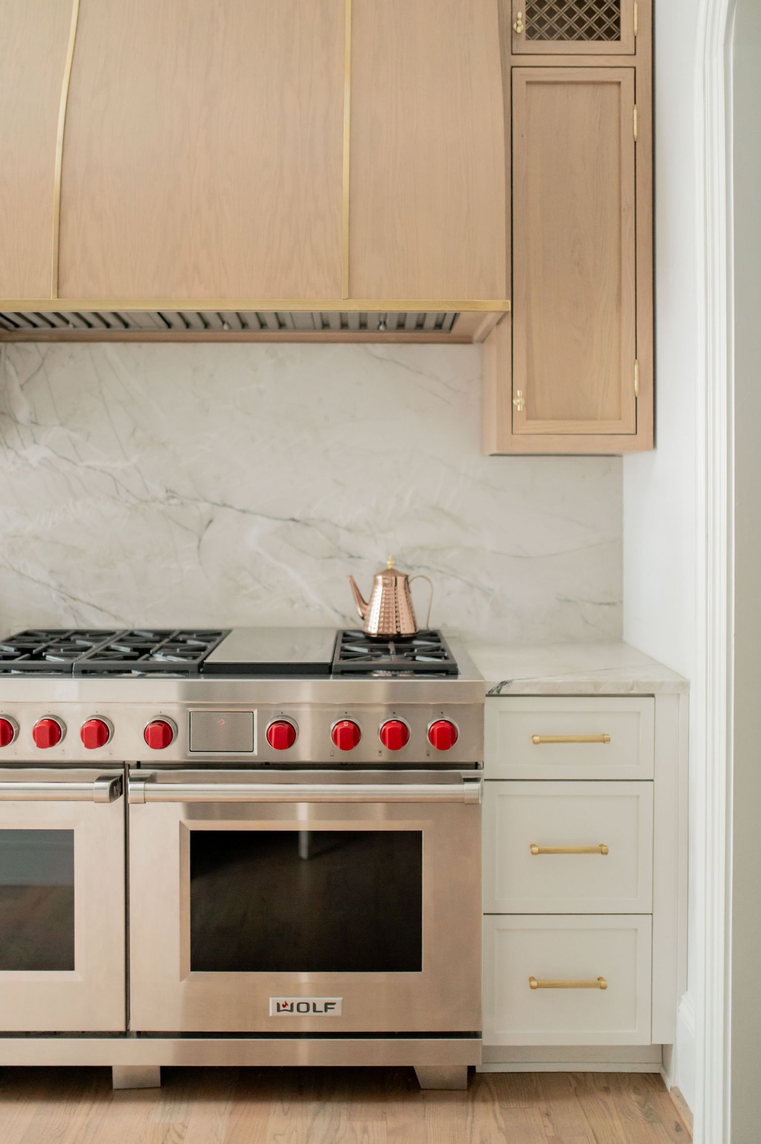 Wolf dual-fuel range with red knobs, marble backsplash, light wood cabinets, and brass hardware in a modern kitchen.