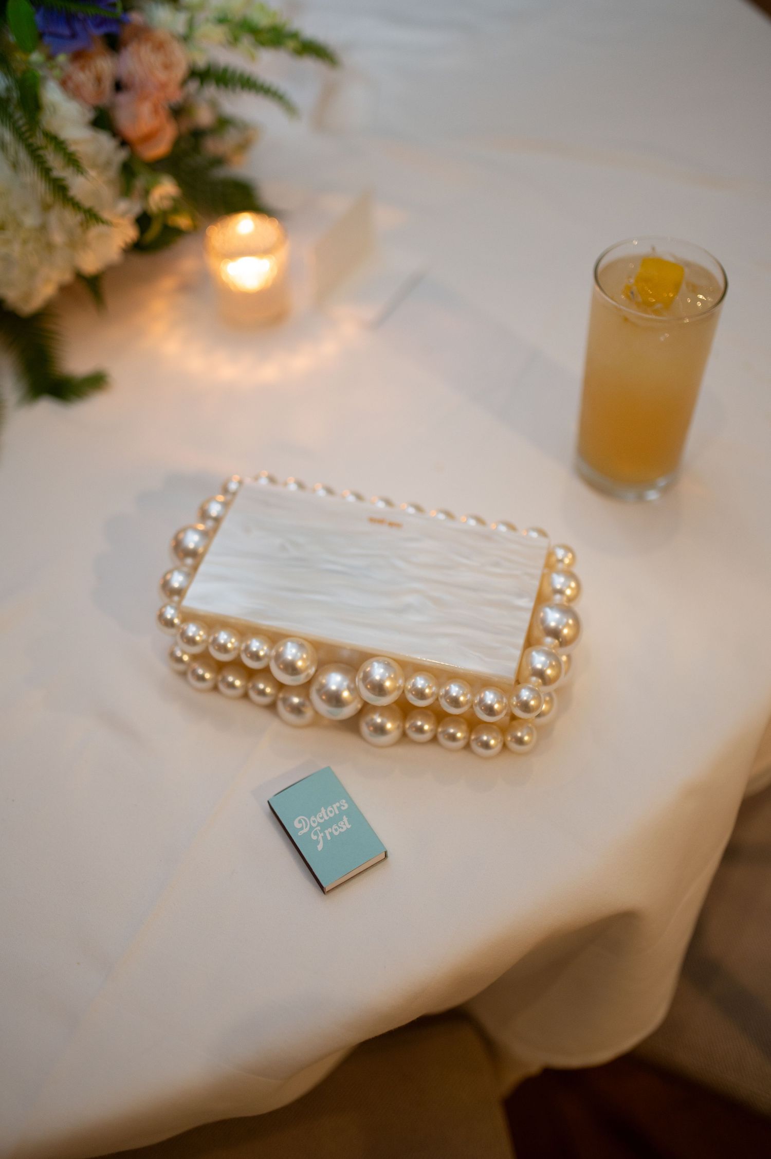 Wedding reception detail with pearl-adorned place card, candle, floral arrangement and cocktail on white tablecloth.