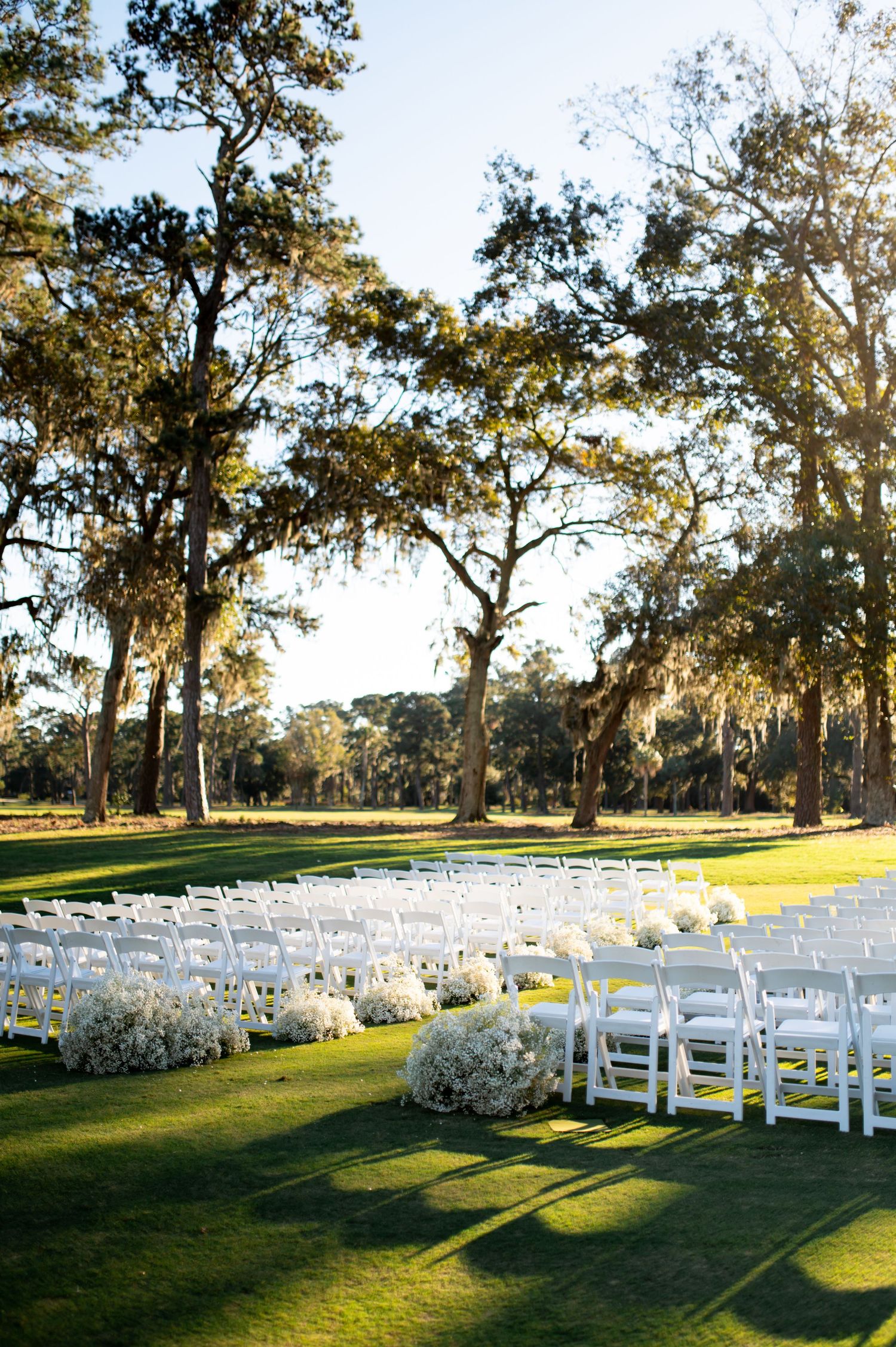 White folding chairs arranged on a grassy lawn under majestic oak trees for an outdoor wedding ceremony.
