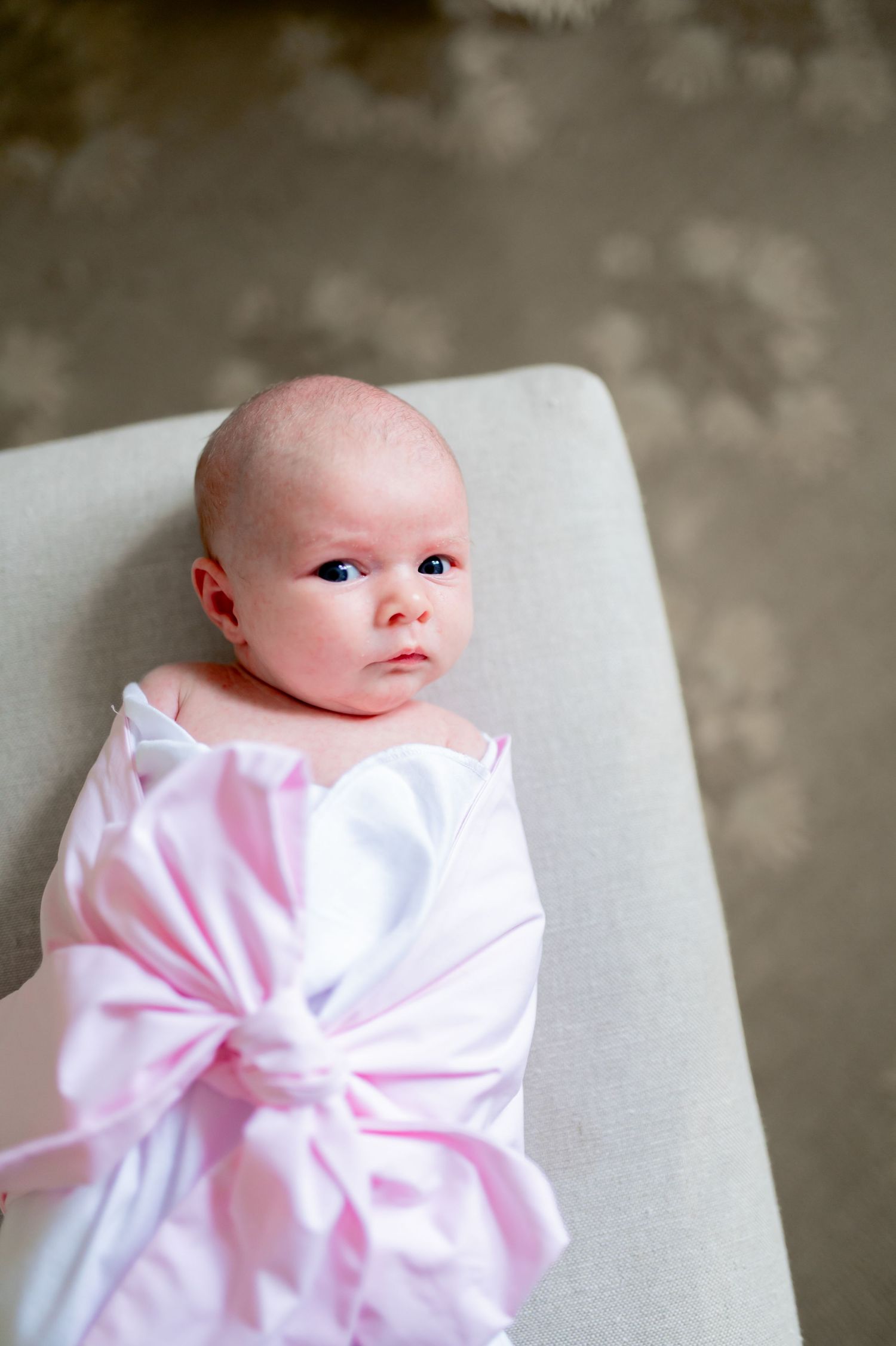 A small baby wrapped in a soft pink blanket lies on a beige surface against a blurred background.