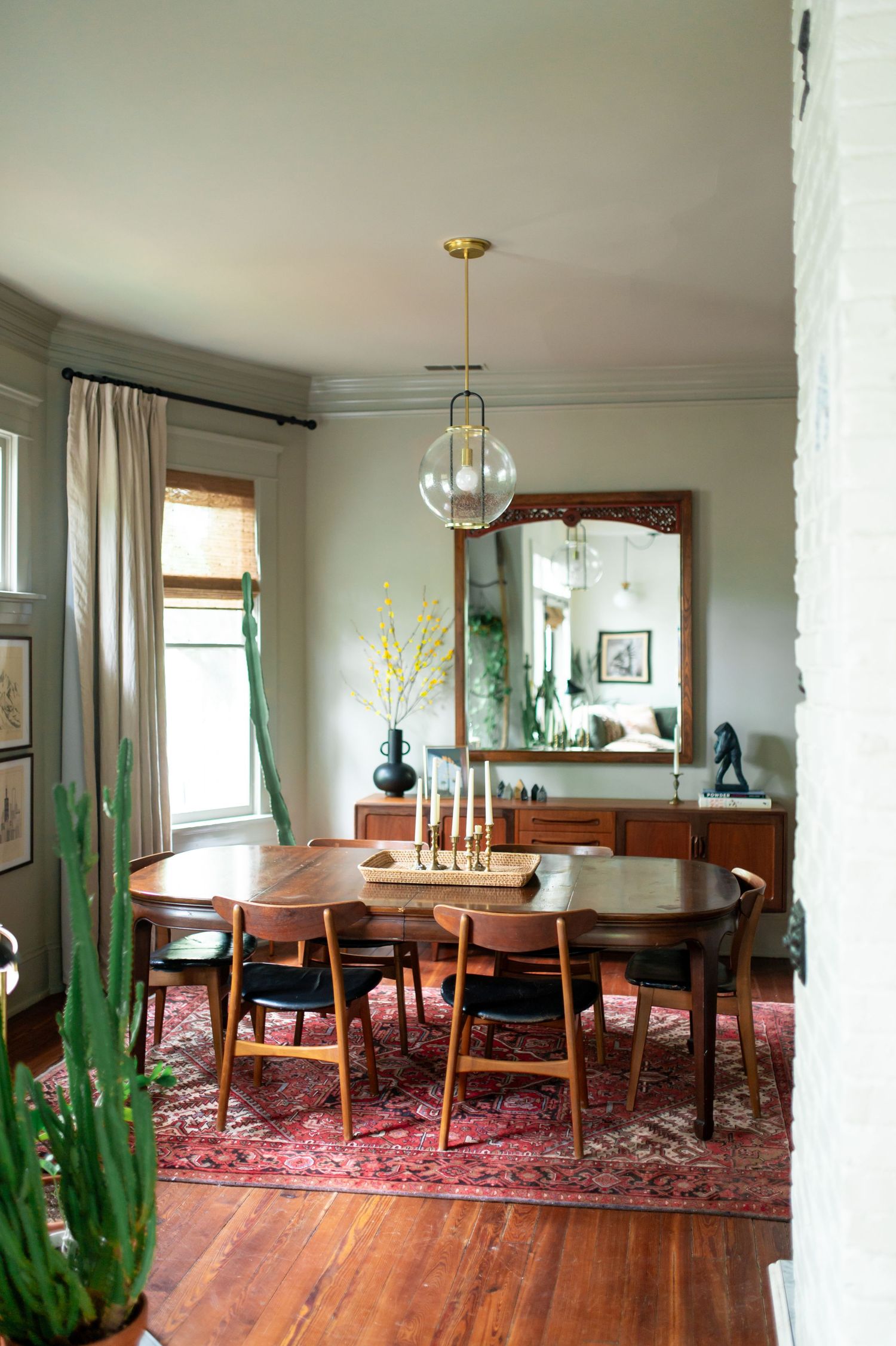 Modern dining room with oval wood table, gold pendant light, red Persian rug, and tall green cactus plants.