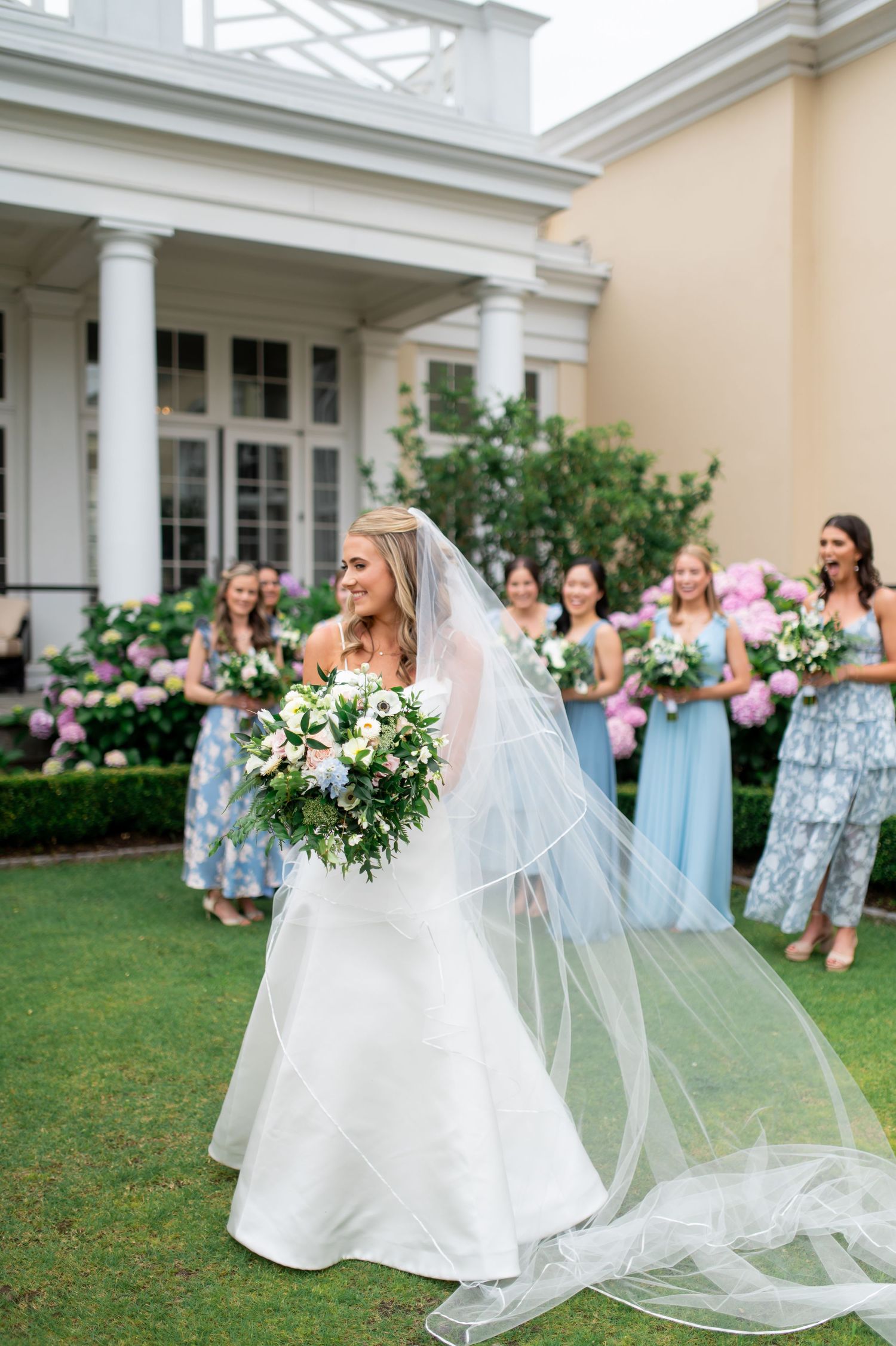 Bride in flowing white gown and veil holds bouquet while bridesmaids stand in background.