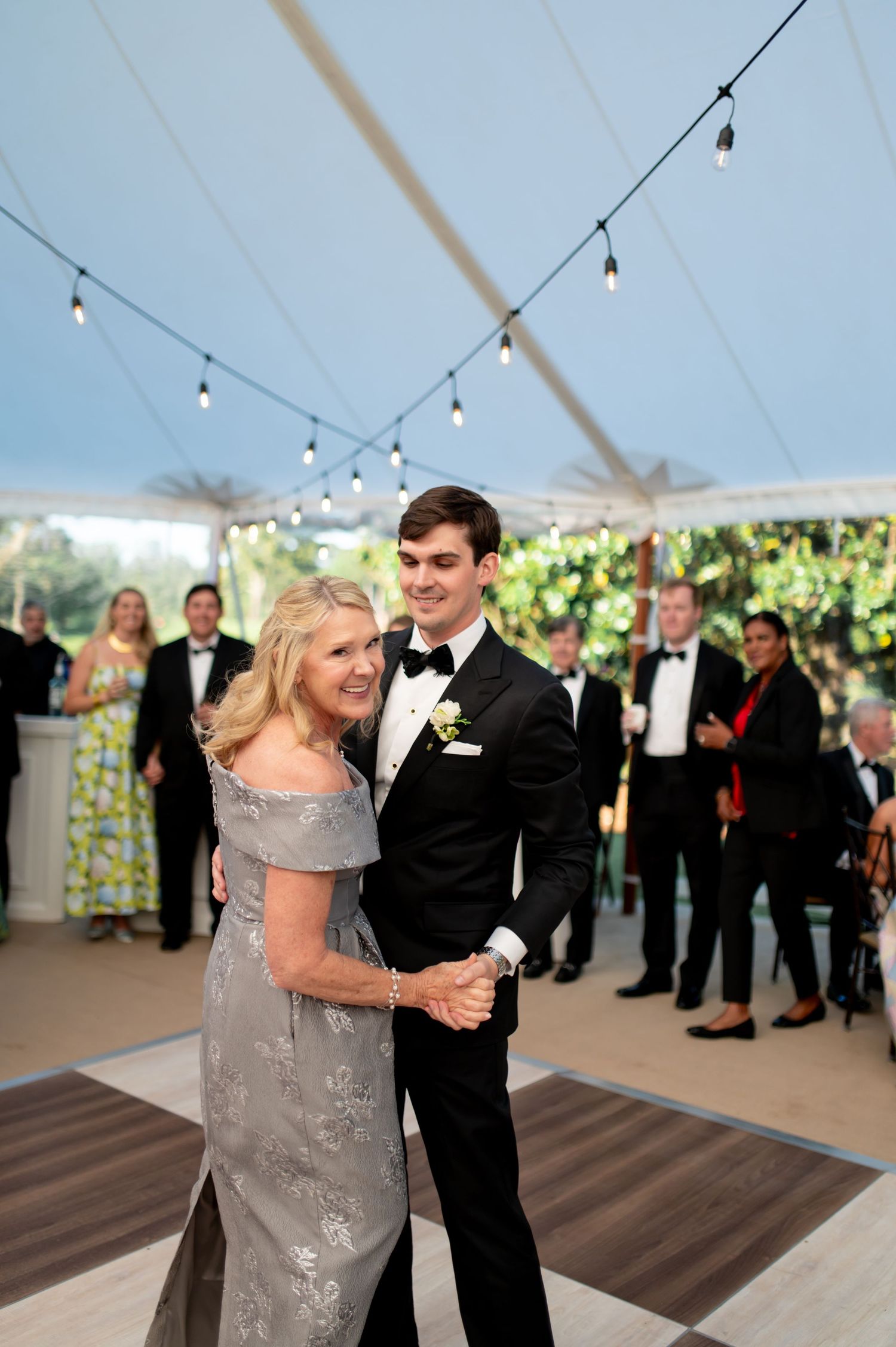 Groom shares a sweet mother-son dance under string lights in a white tent at wedding reception.