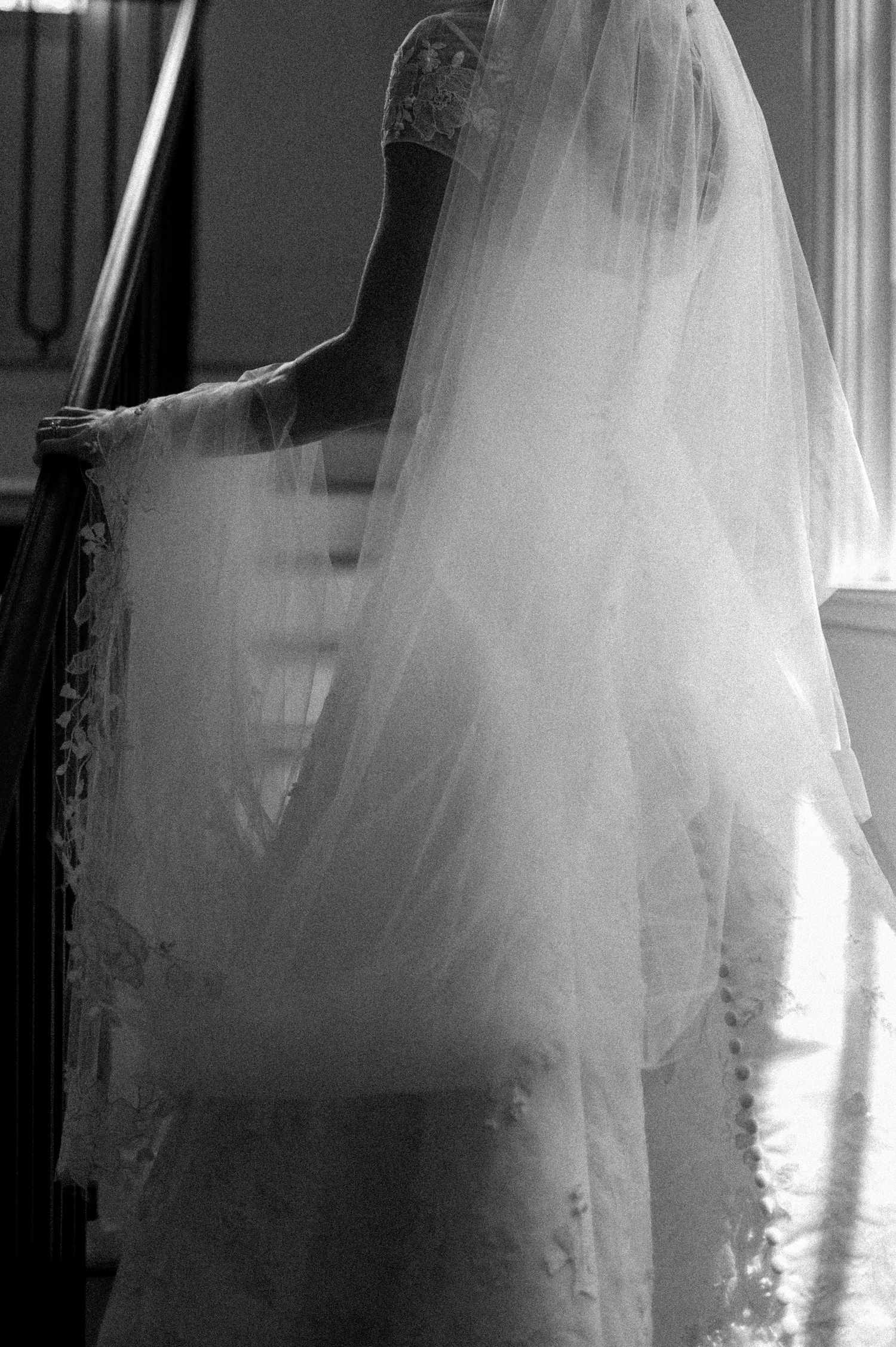 Black and white photo of bride in lace wedding gown and long veil descending staircase.