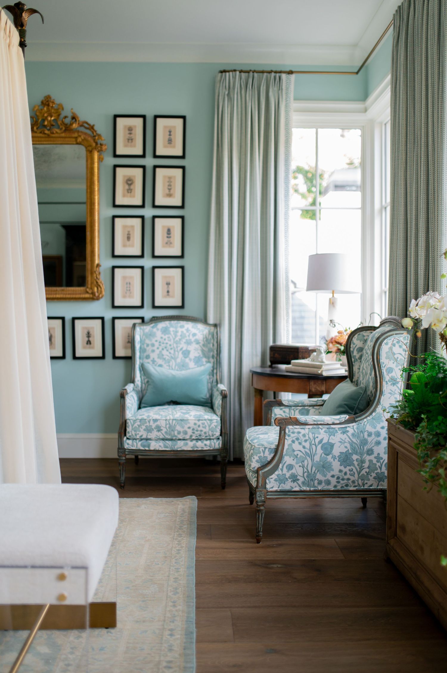 Elegant bedroom with seafoam green walls, ornate gold mirror, white curtains and blue floral chaise lounge on dark hardwood floors.