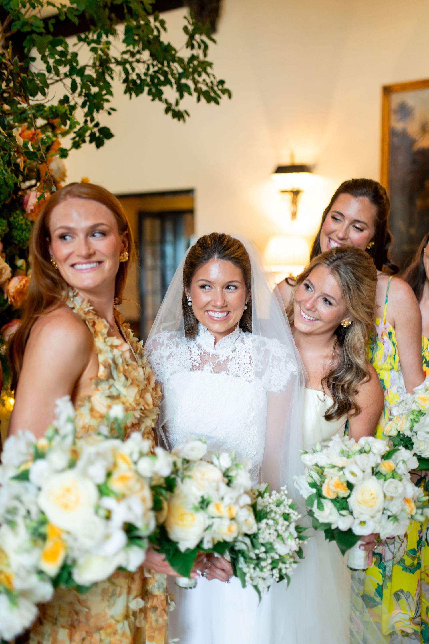 Wedding party members smile and laugh while holding white floral bouquets in an indoor venue.