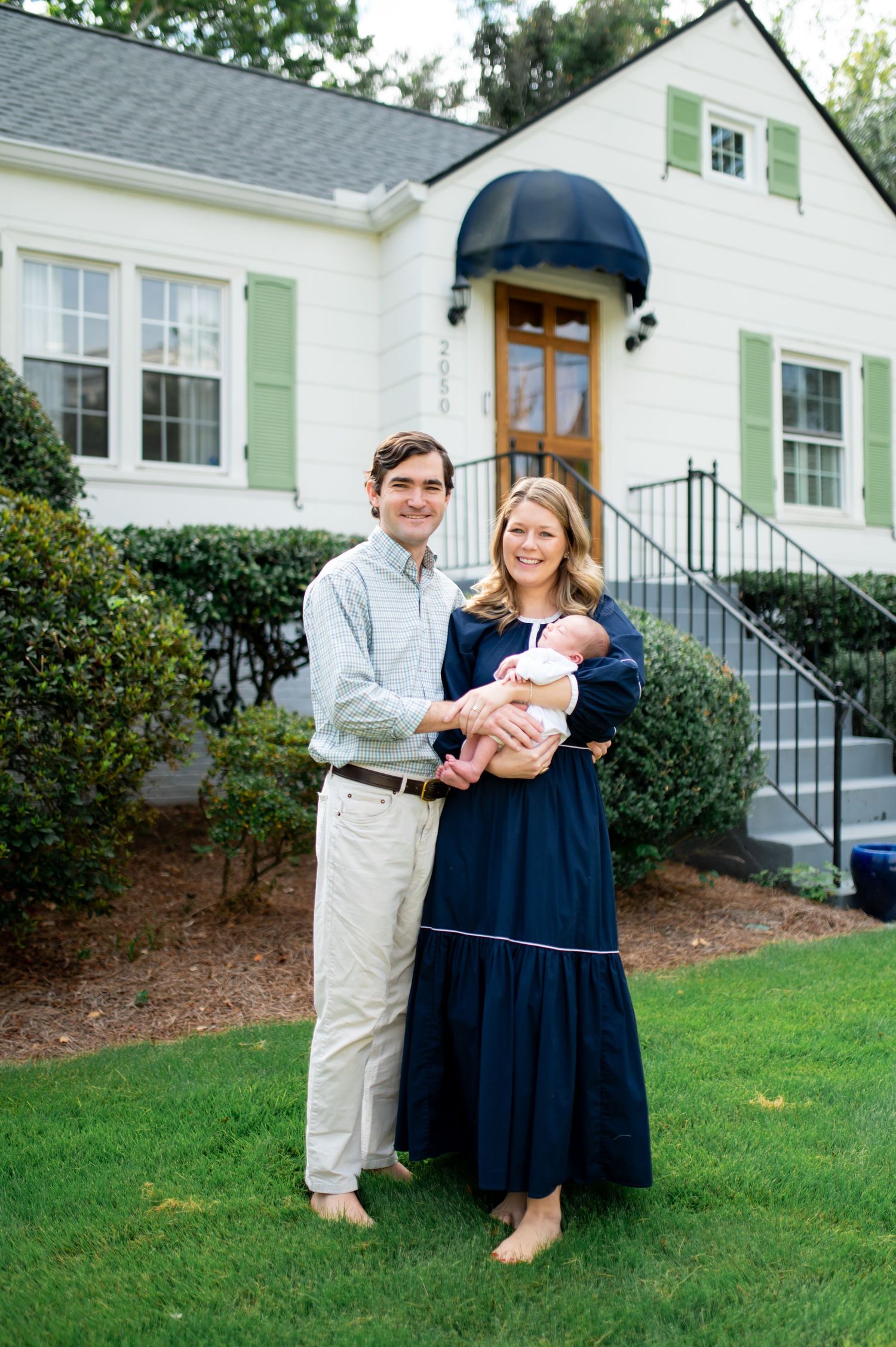 A family poses together on the lawn in front of a white house with green shutters.