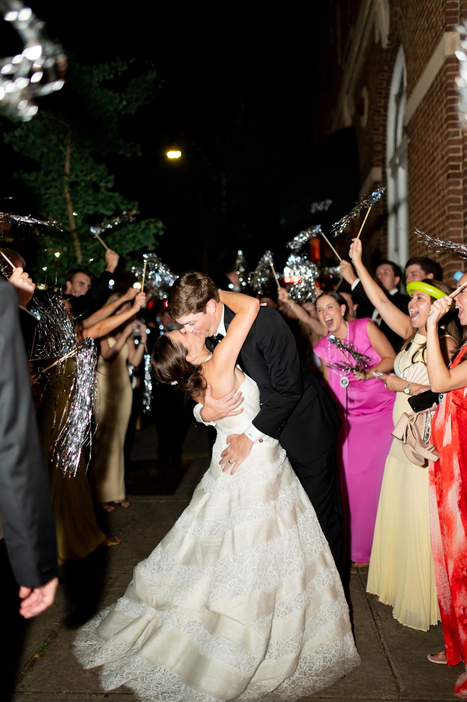 Groom dips bride for romantic kiss during sparkler and tinsel wand sendoff on dark evening street.